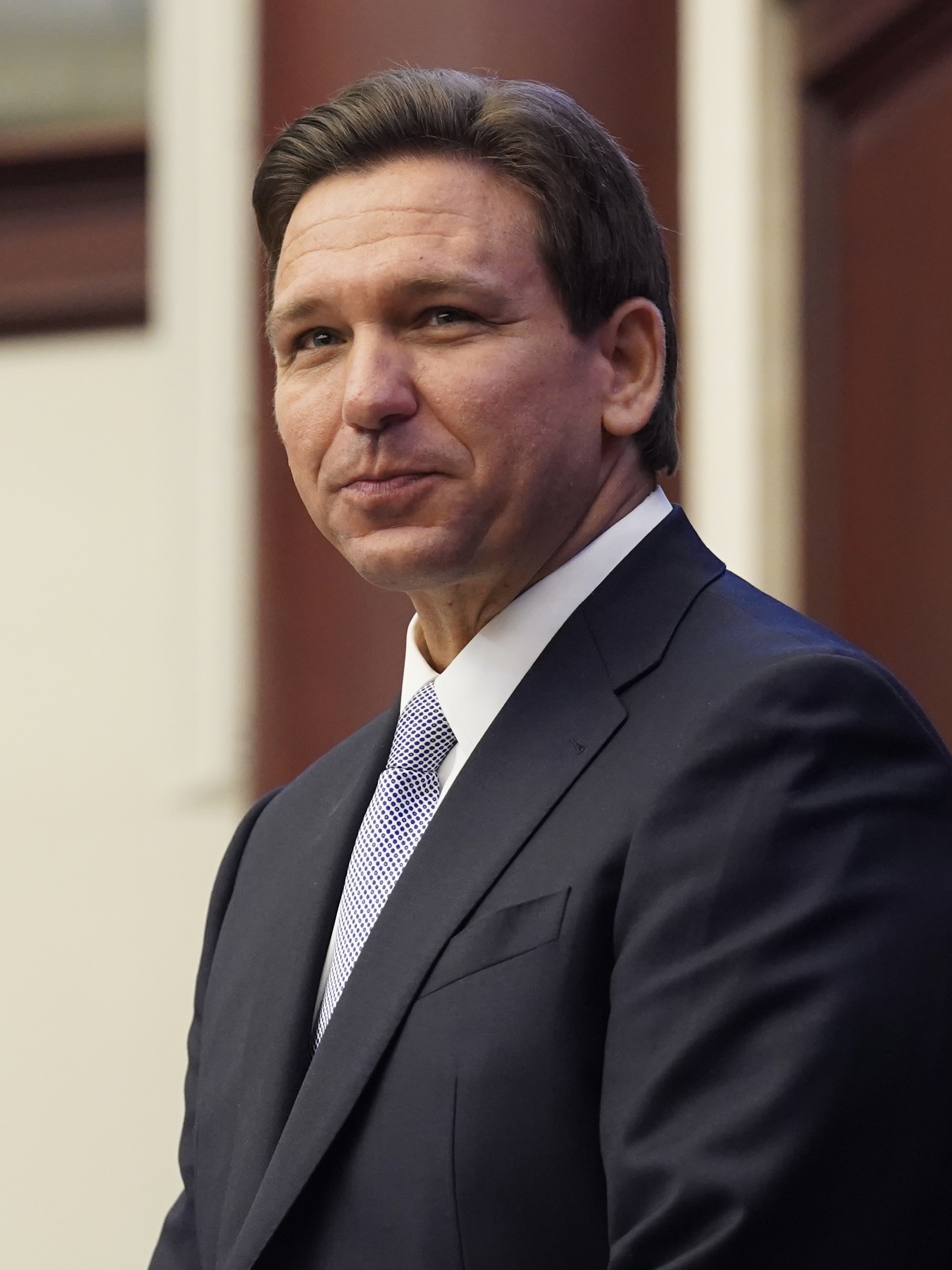 Man in dark suit and blue tie speaking at a podium with white columns behind him.