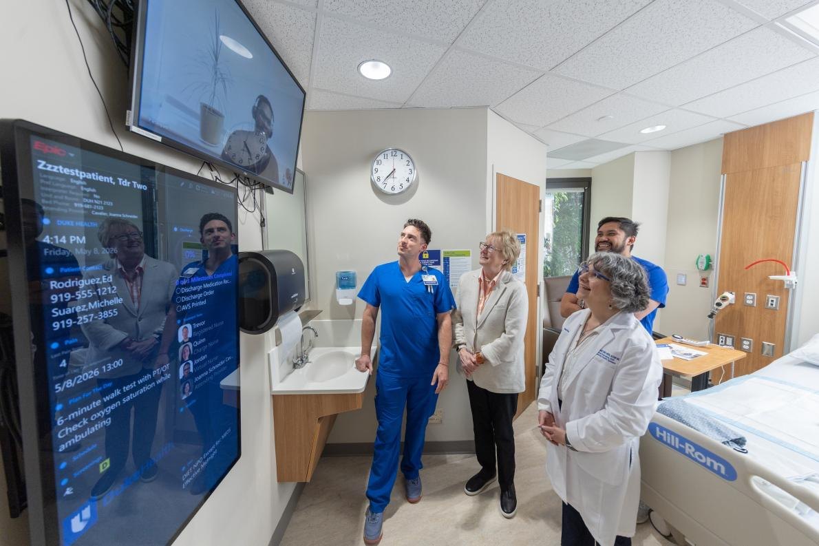 Five people standing in a modern hospital room with a large digital display screen and medical equipment.