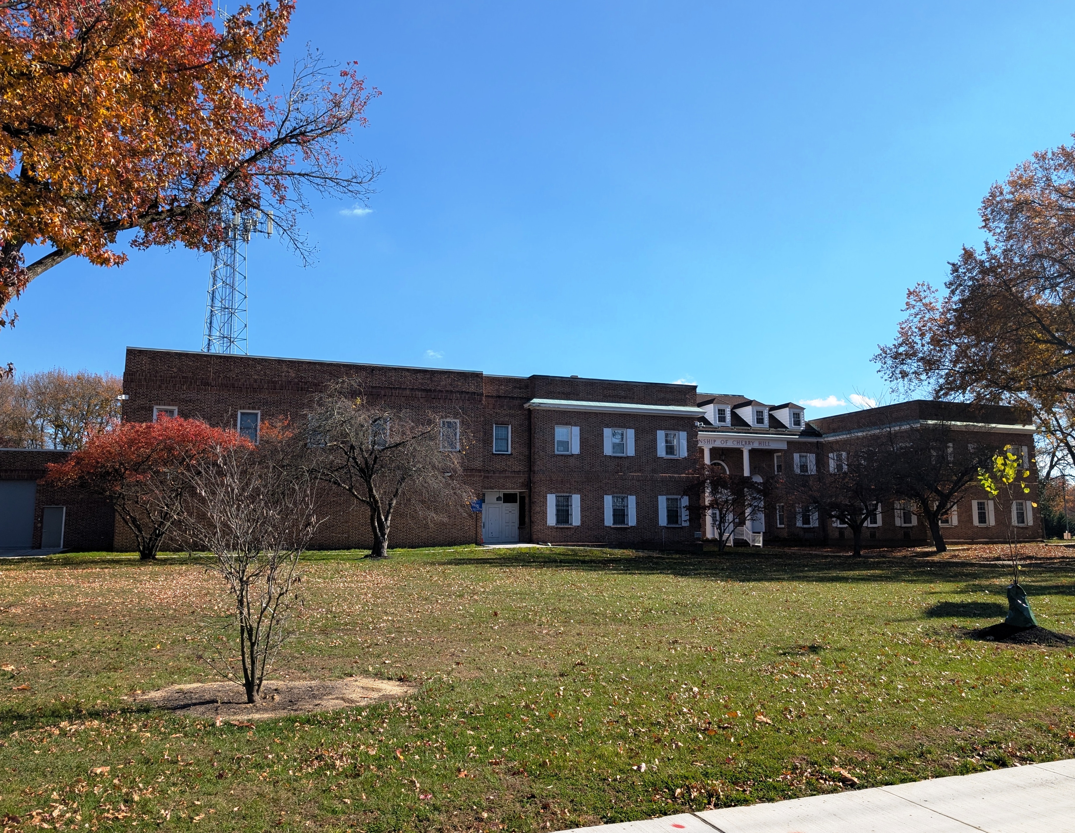 Two-story red-brick municipal building surrounded by autumn trees and fallen leaves.
