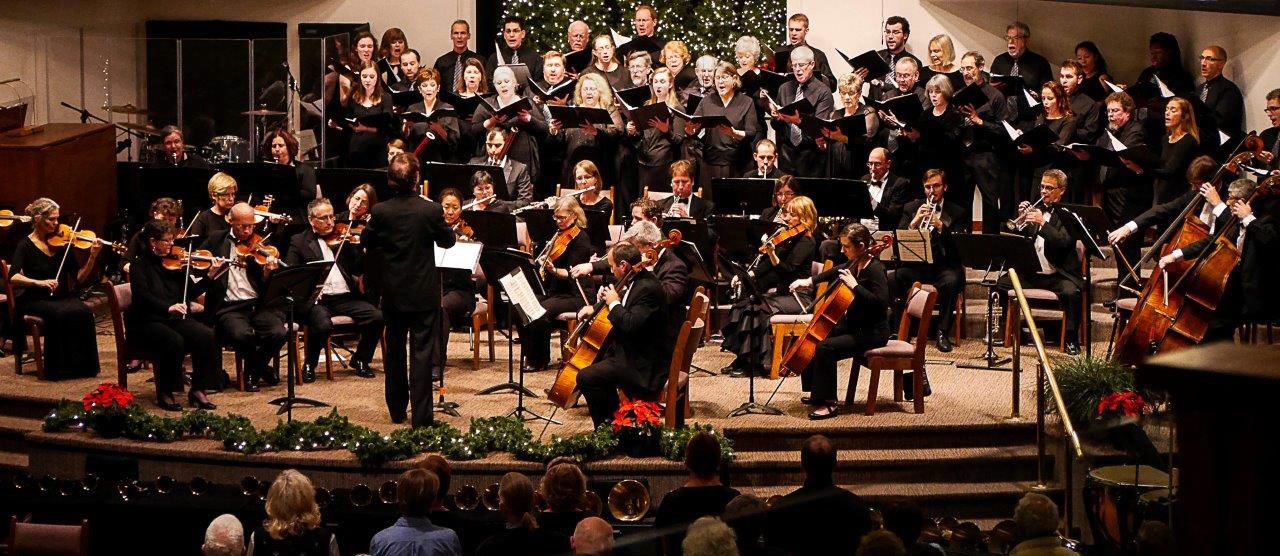 Symphony orchestra and choir performing on a stage decorated with Christmas trees and holiday greenery.