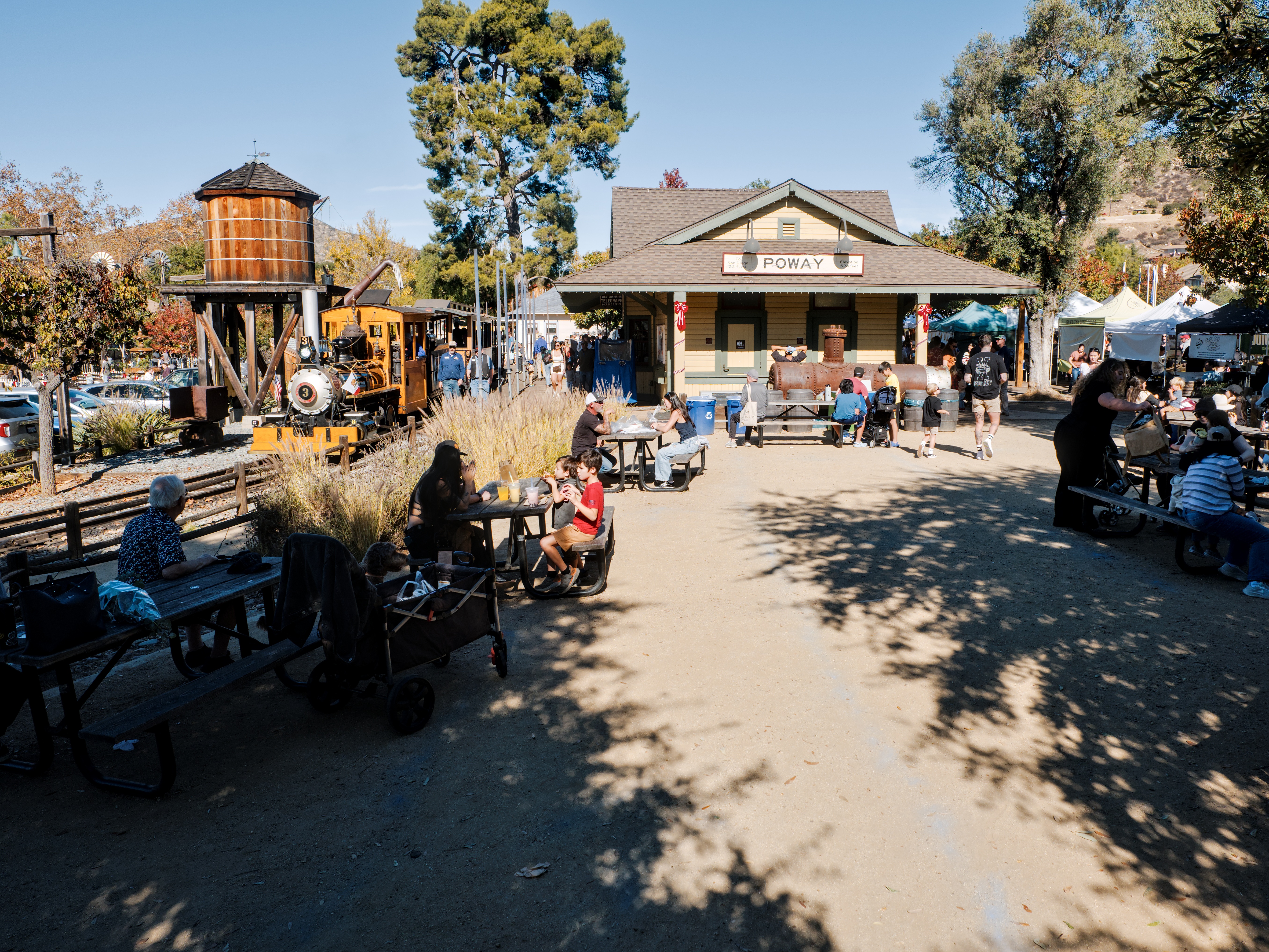 Outdoor park gathering area with historic depot building, vintage locomotive, and picnic tables under autumn trees.