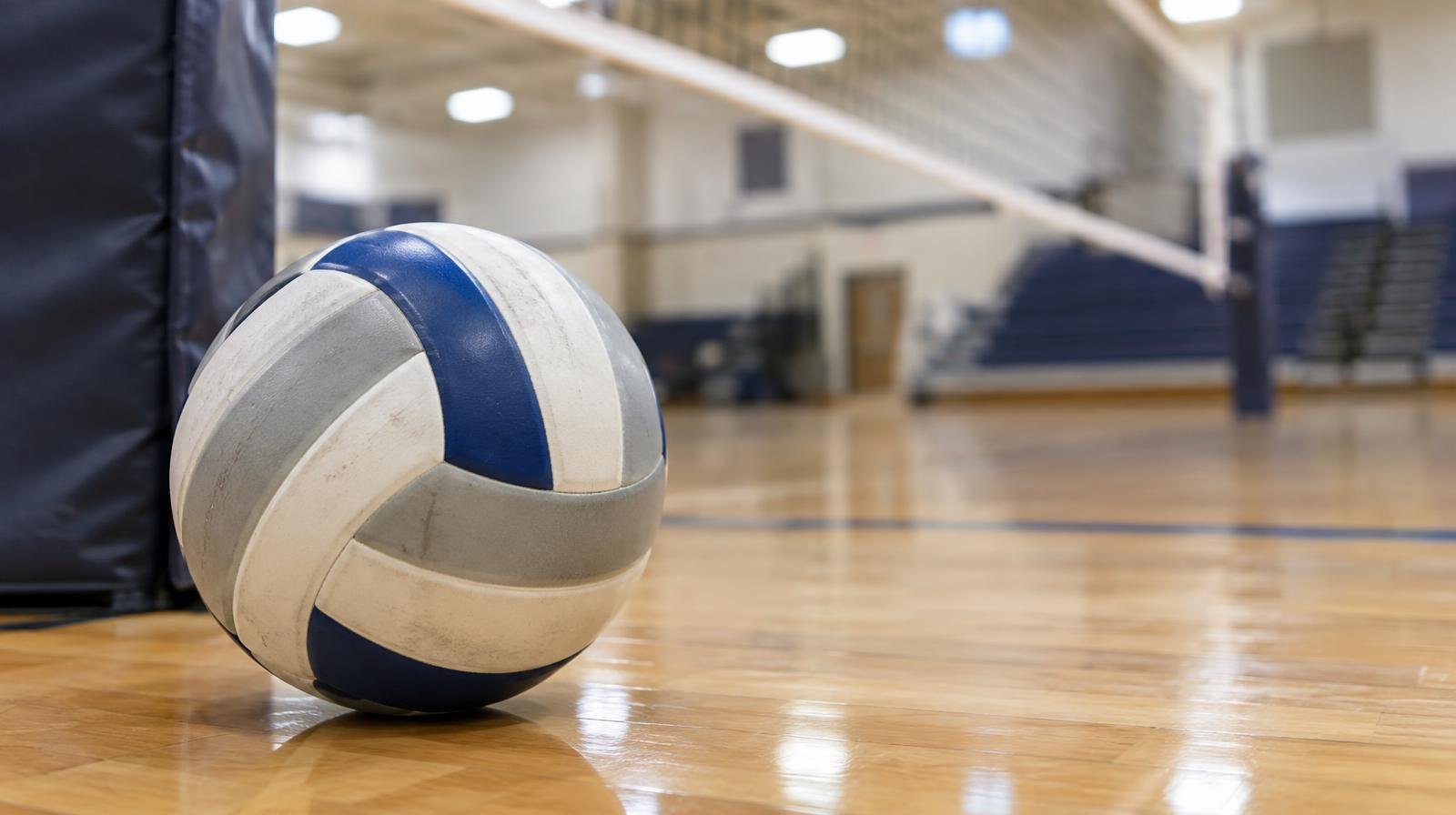 A volleyball resting on a hardwood gymnasium floor near a net post.