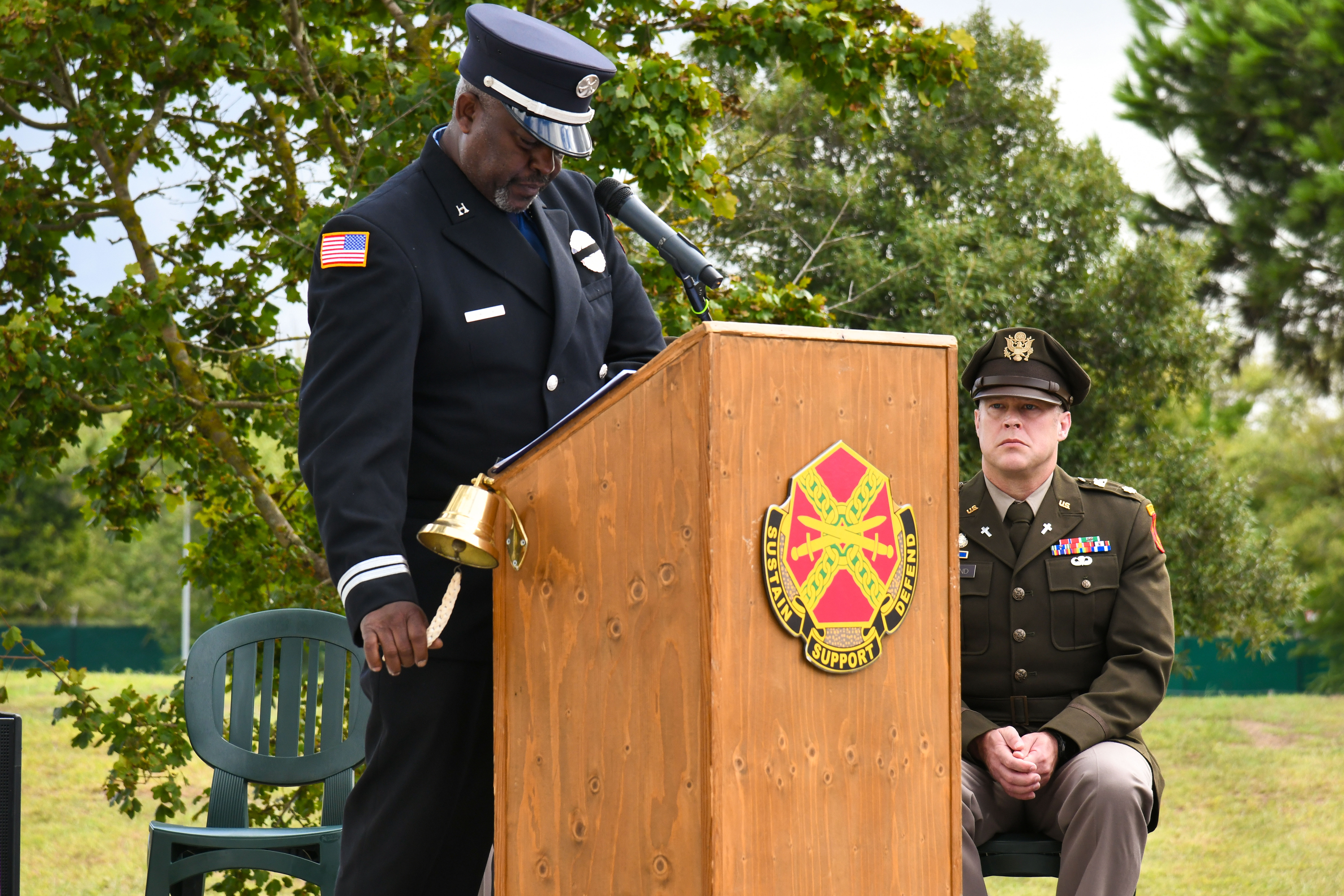 Firefighter in dress uniform rings a bell at a podium during an outdoor memorial ceremony.