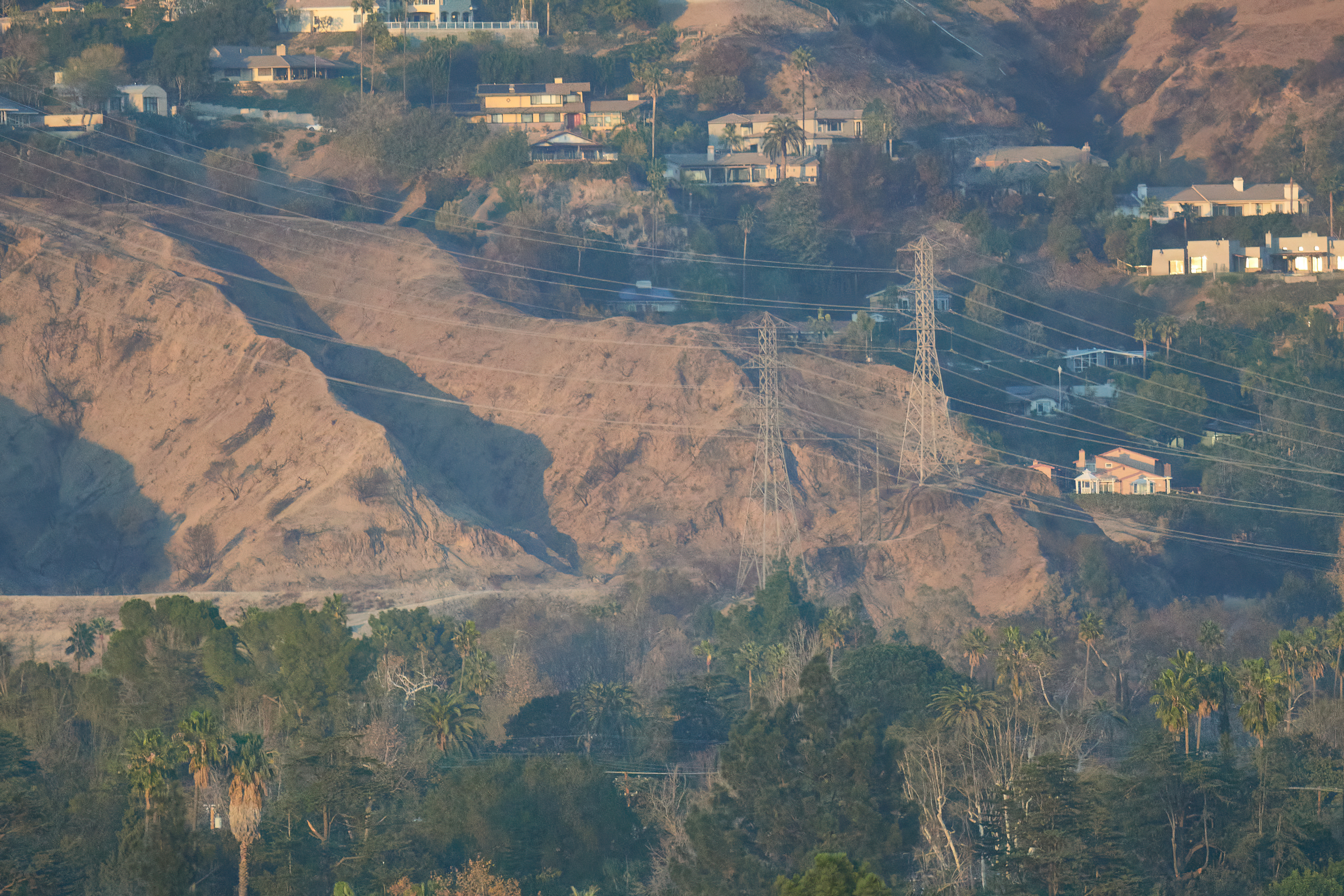 Burned hillsides with exposed soil, a transmission tower, and homes on a ridgeline near a wash.