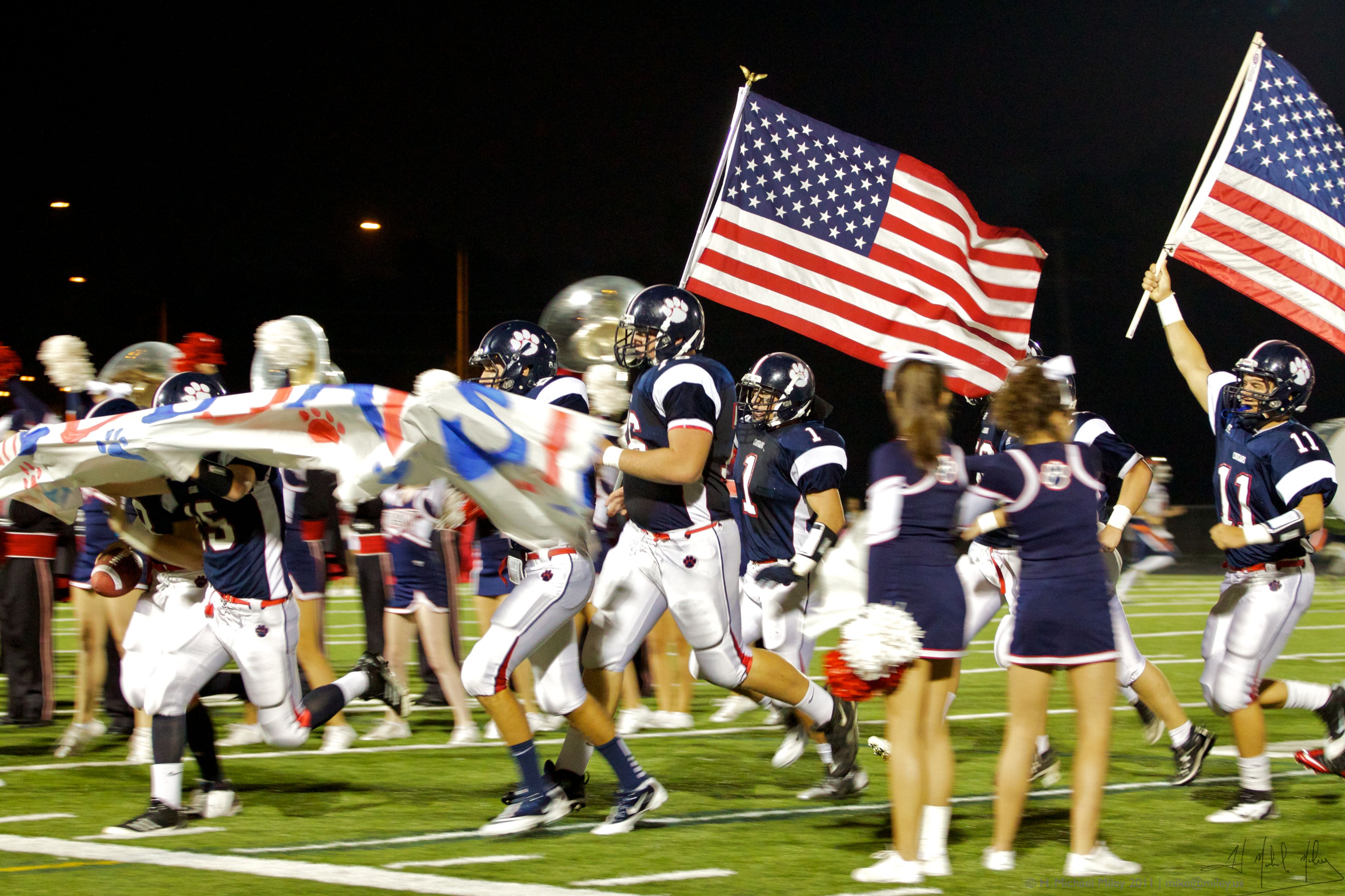 High school football players and cheerleaders running onto a lit field at night carrying American flags.
