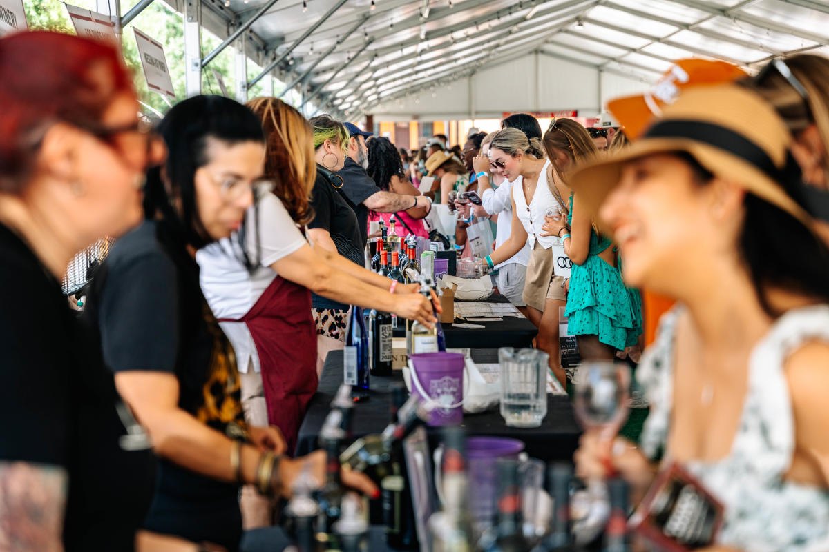 A large crowd of people sampling wines along a long table under a white tent.