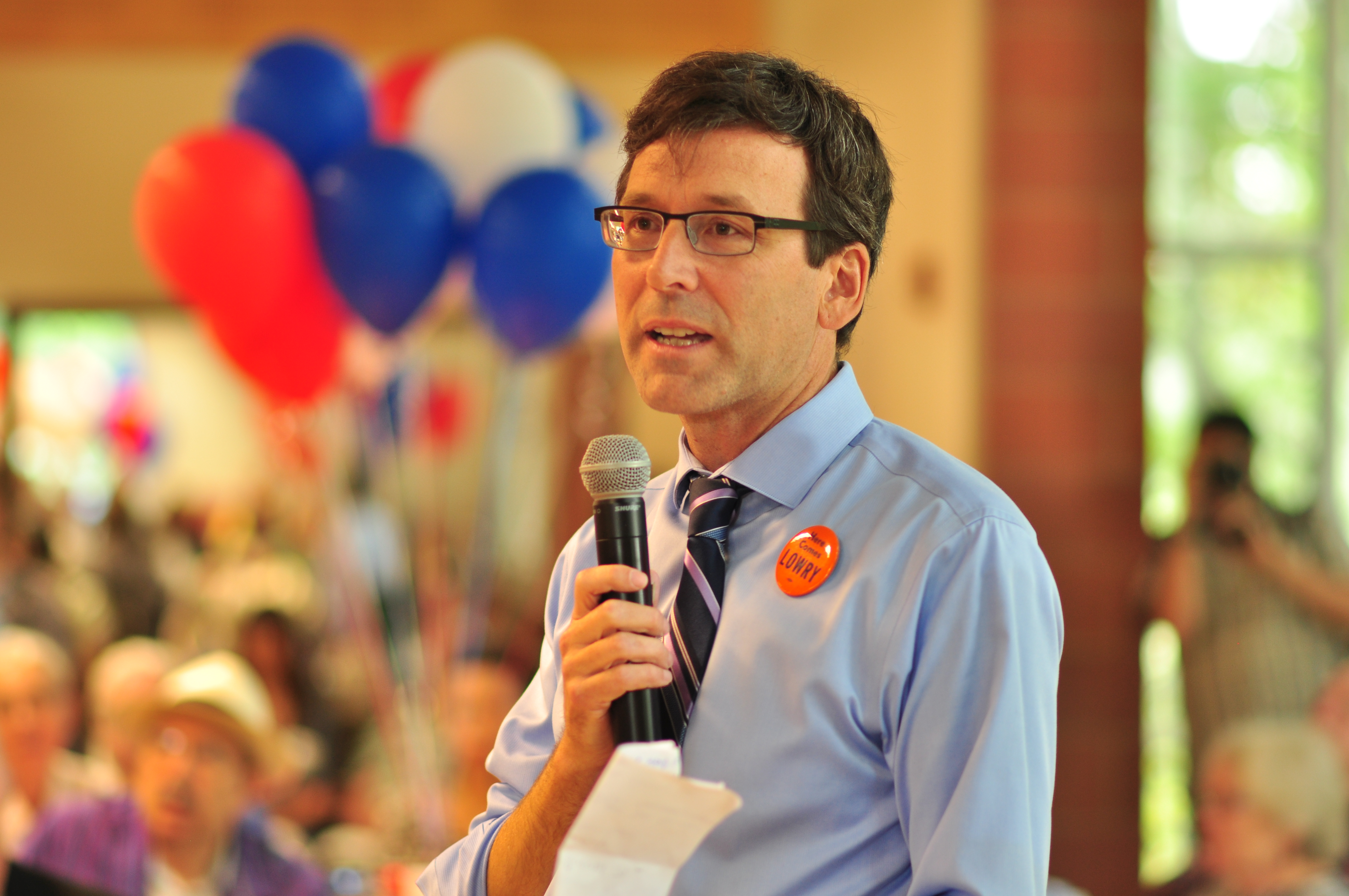 Man in glasses and blue dress shirt speaking into a microphone at an indoor event with red, white, and blue balloons.