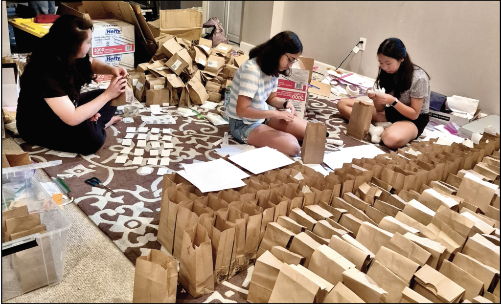 Three young women sit on a rug packaging rows of brown paper bags with supplies.