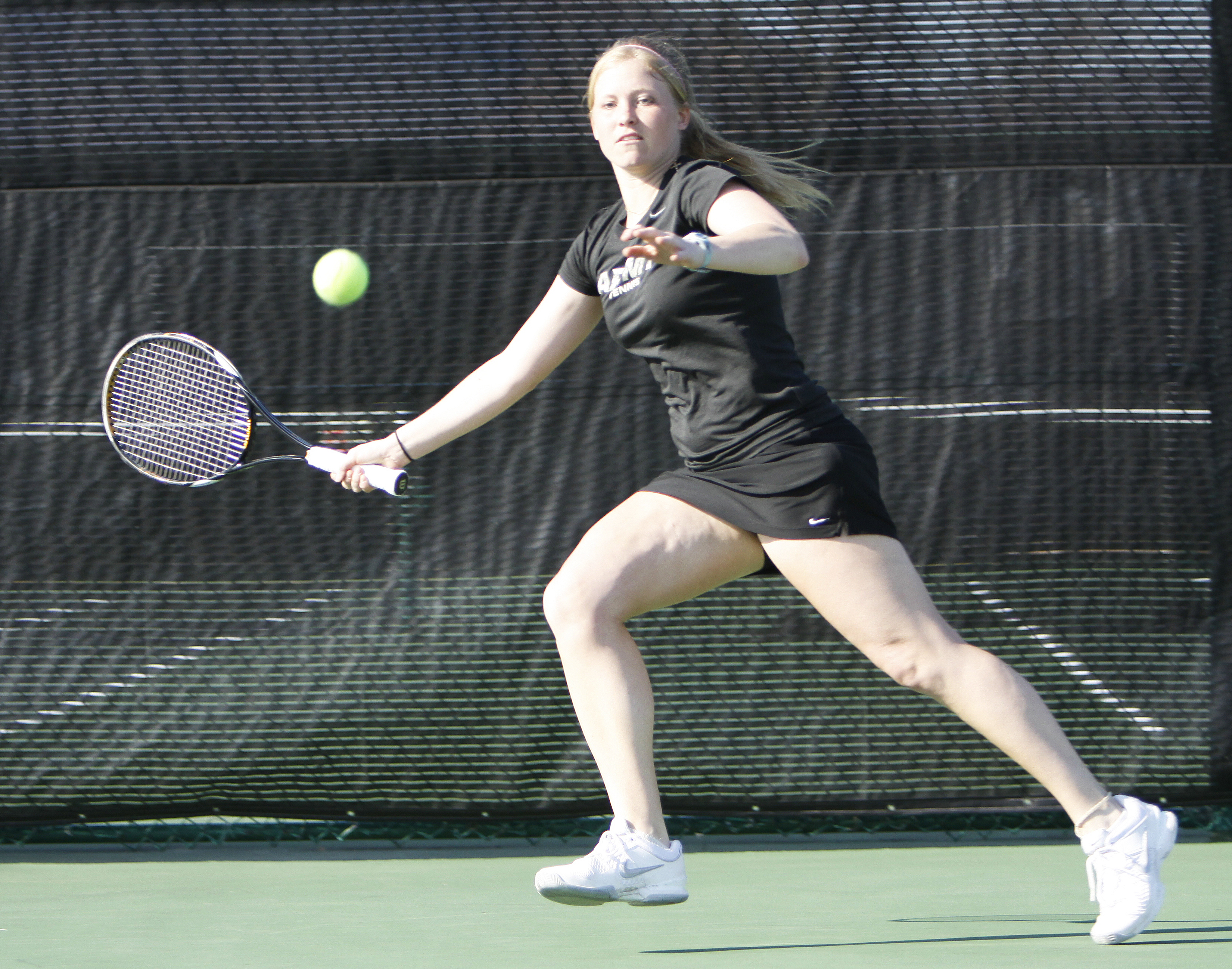 Female tennis player in black outfit lunges to hit a forehand on an outdoor court.