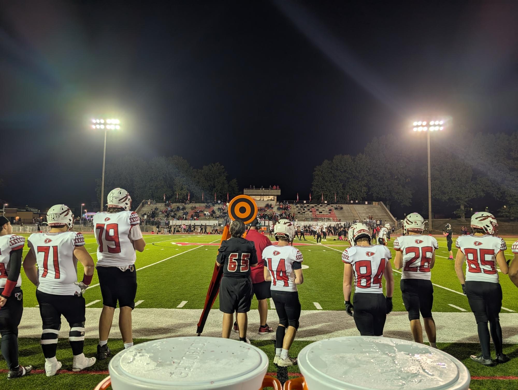 Football players in red and white uniforms stand on a sideline under stadium lights at night.