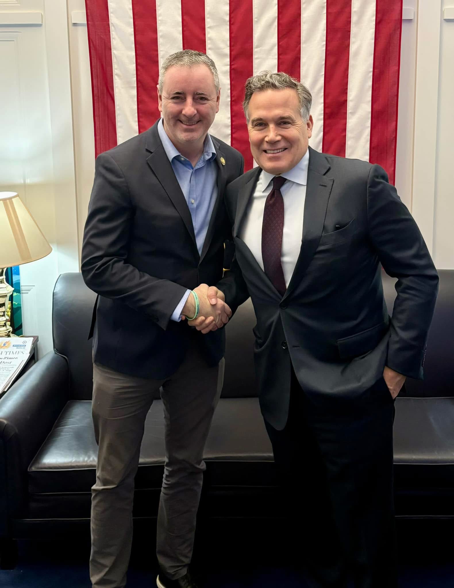 Two men in dark suits shake hands in front of a large American flag indoors.