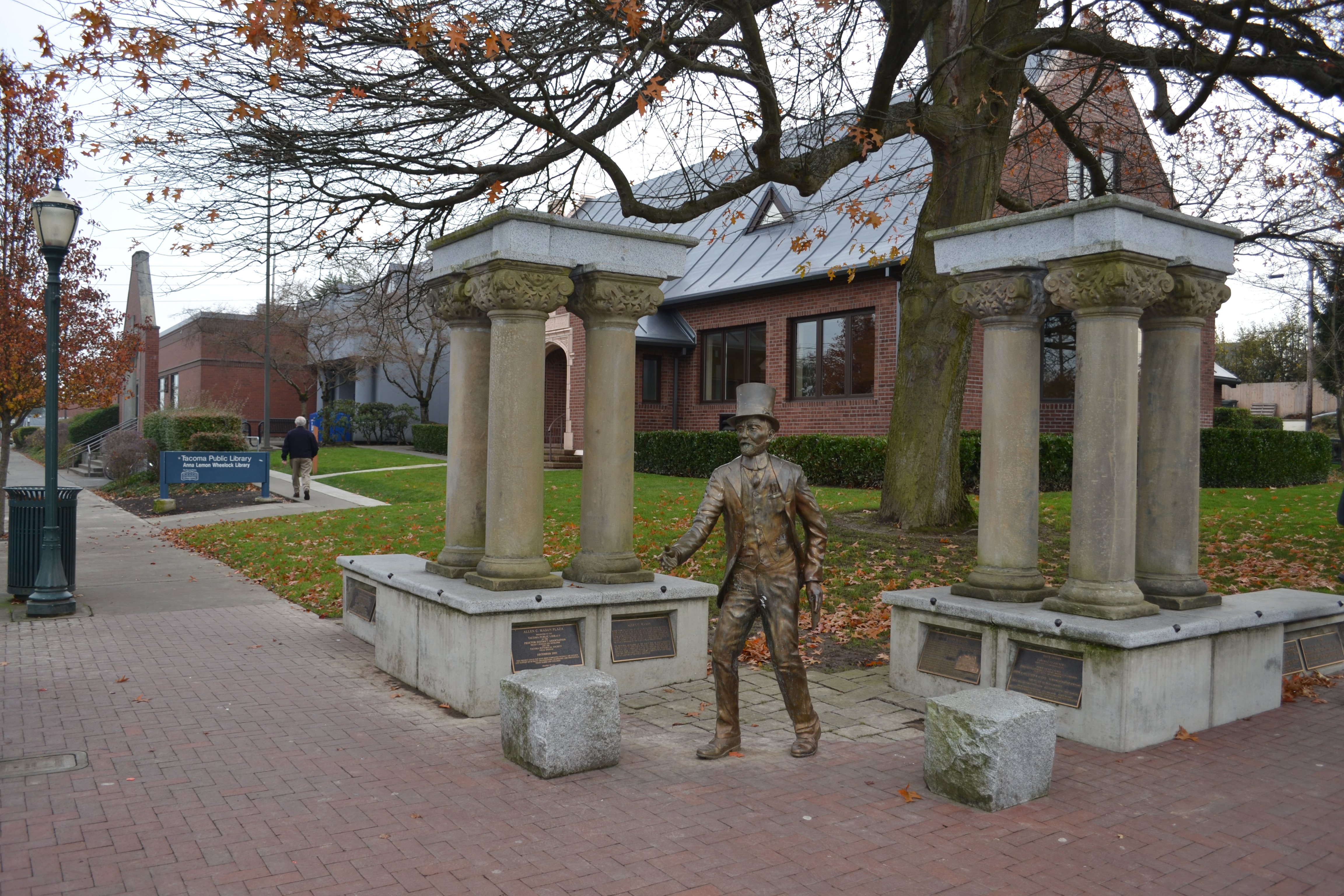 Bronze statue between stone columns outside a brick public library building in autumn.