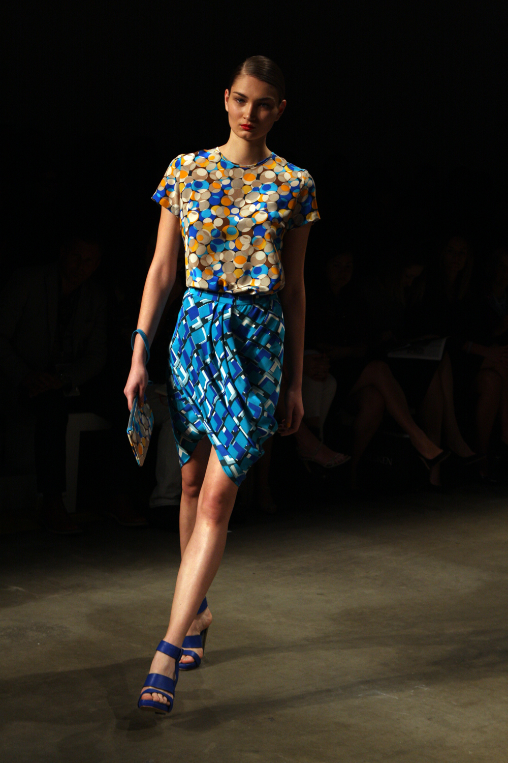 A model in colorful patterned garments walks a dimly lit fashion runway.