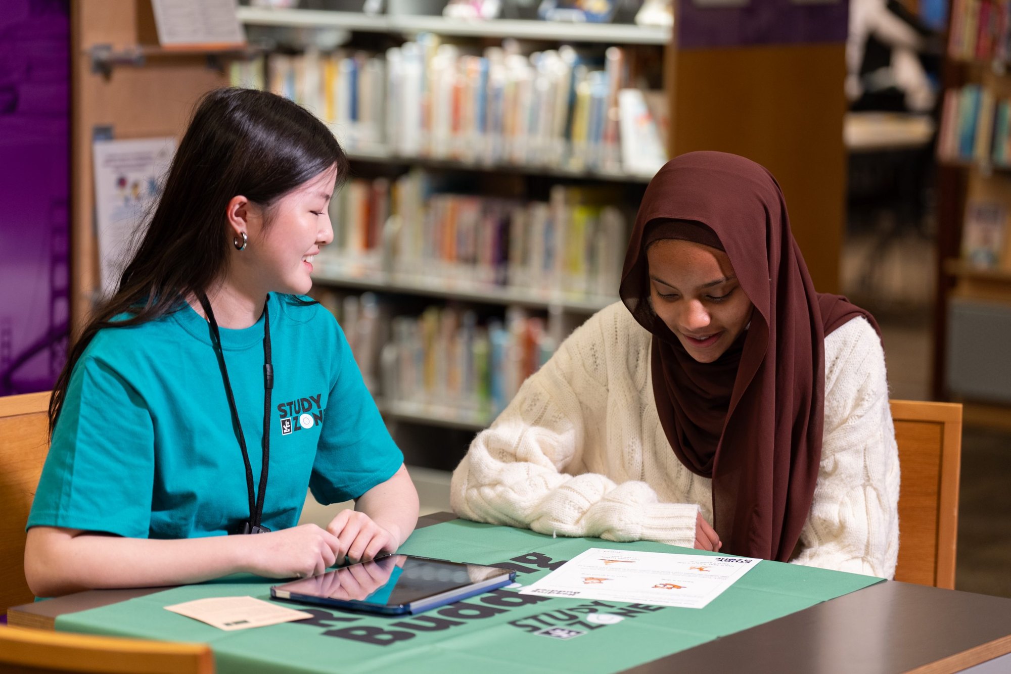 Two people sit at a library table reviewing papers, one wearing a teal shirt and lanyard.