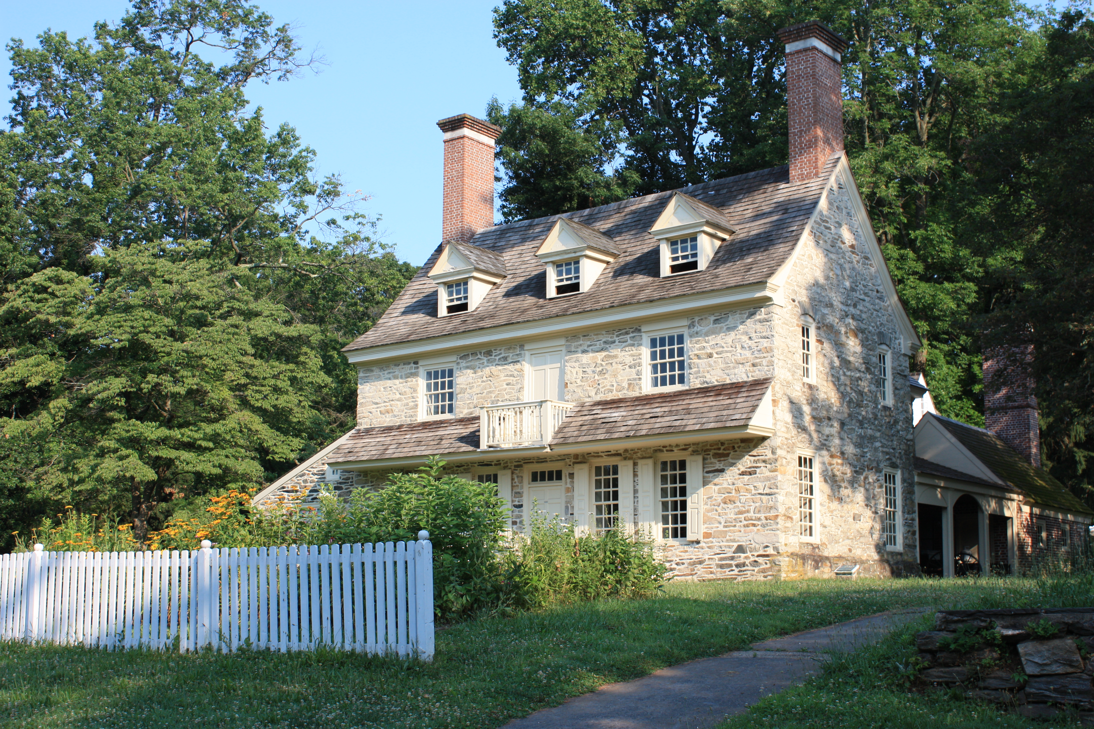 A historic stone farmhouse with dormer windows and a white picket fence on a sunny day.