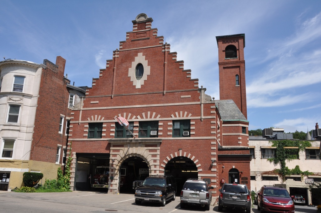 A historic red brick fire station with a stepped gable facade, arched entrances, and a bell tower.