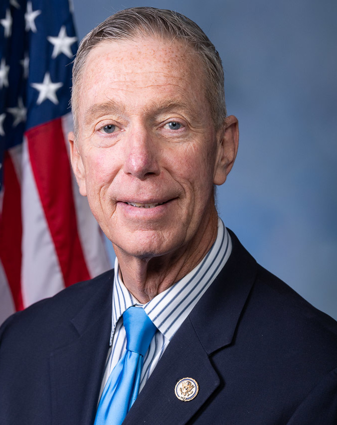 Man in dark suit and blue tie with congressional lapel pin, posed before an American flag.