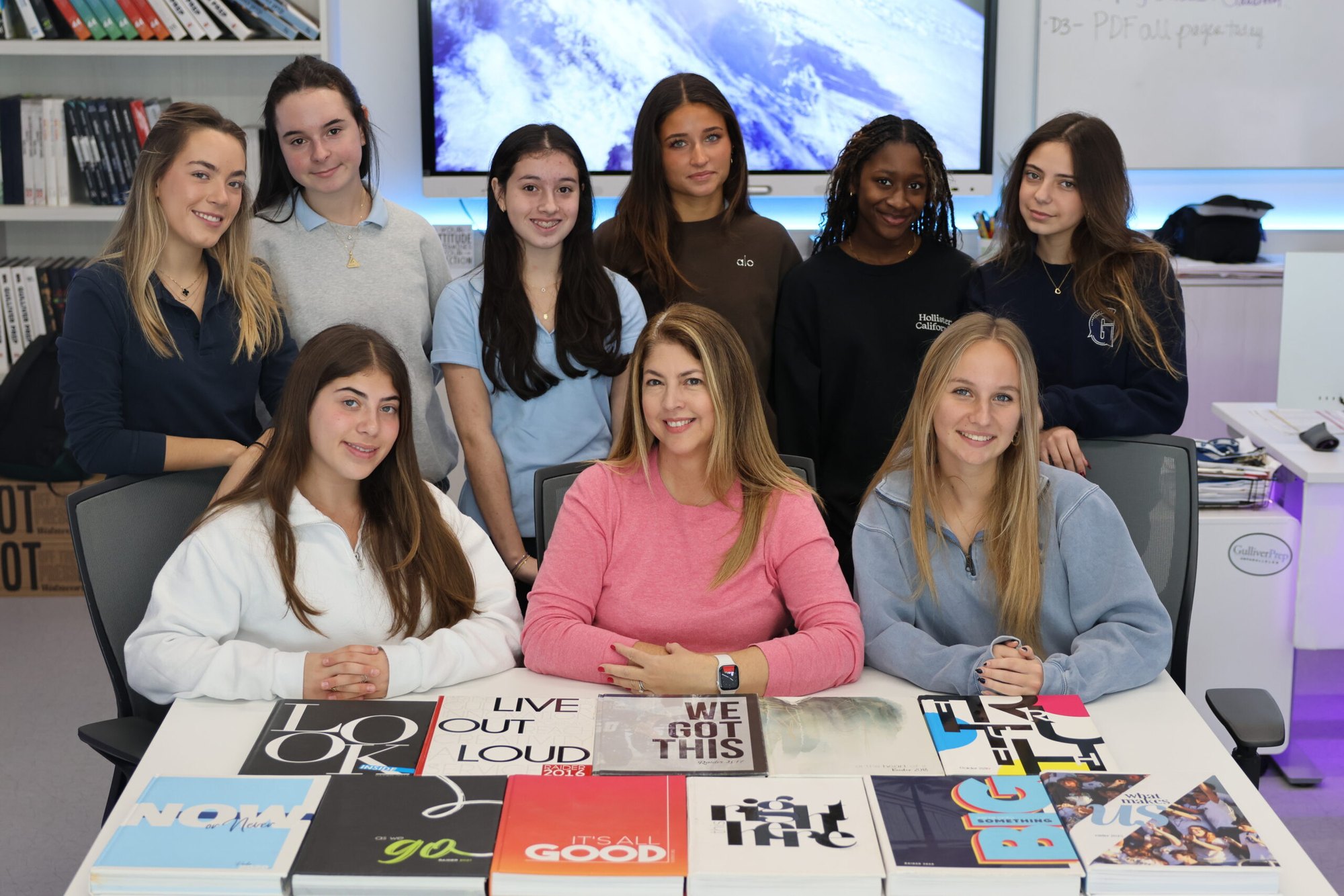 Students and an adult adviser seated at a table covered with printed publications in a school media room.