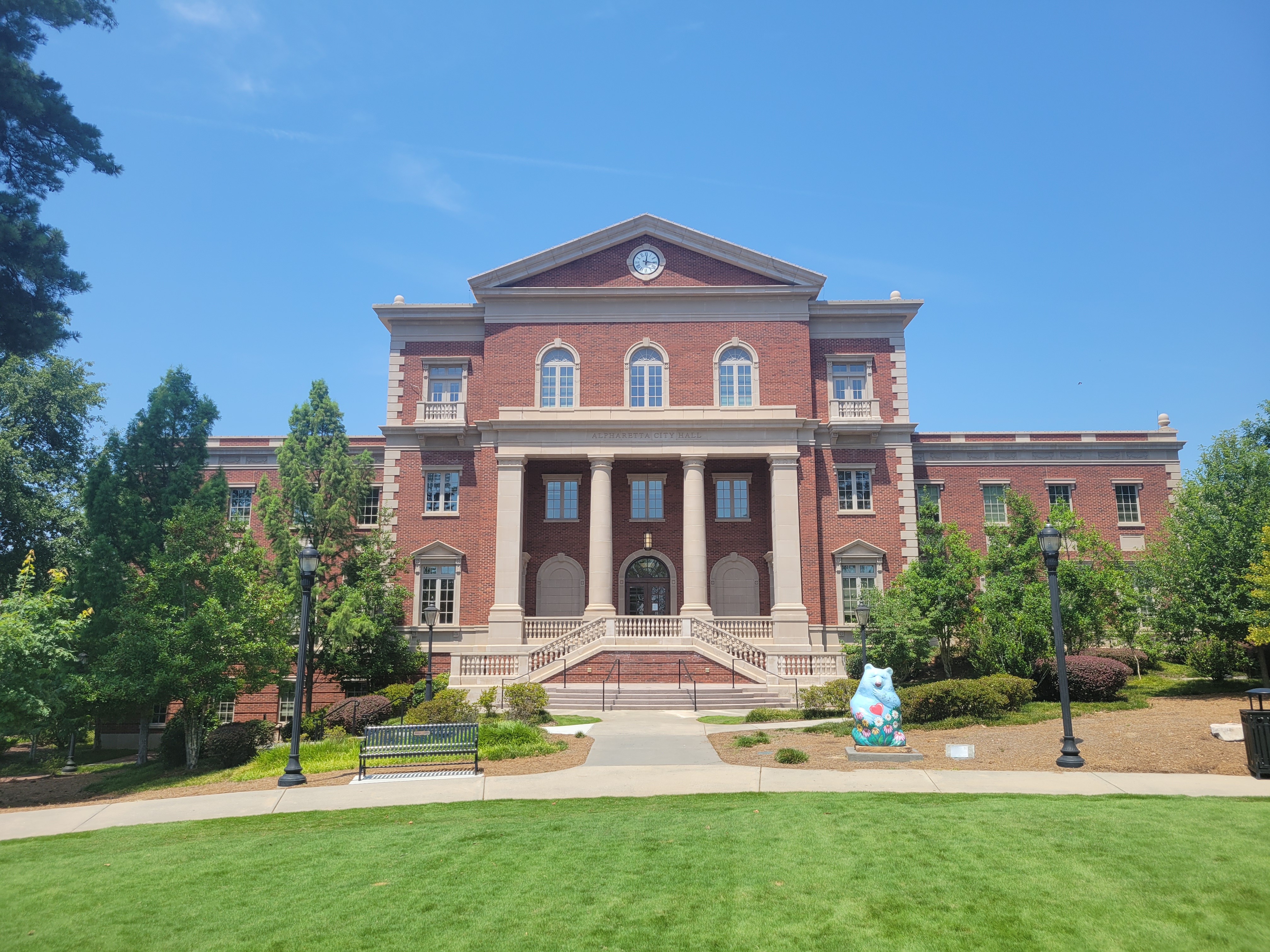 Neoclassical red-brick government building with white columns, green lawn, and a decorative sculpture.