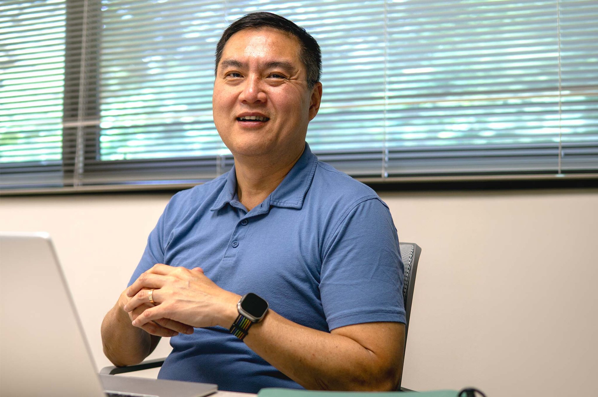 A man in a blue polo shirt sits smiling at a desk in an office with window blinds behind him.