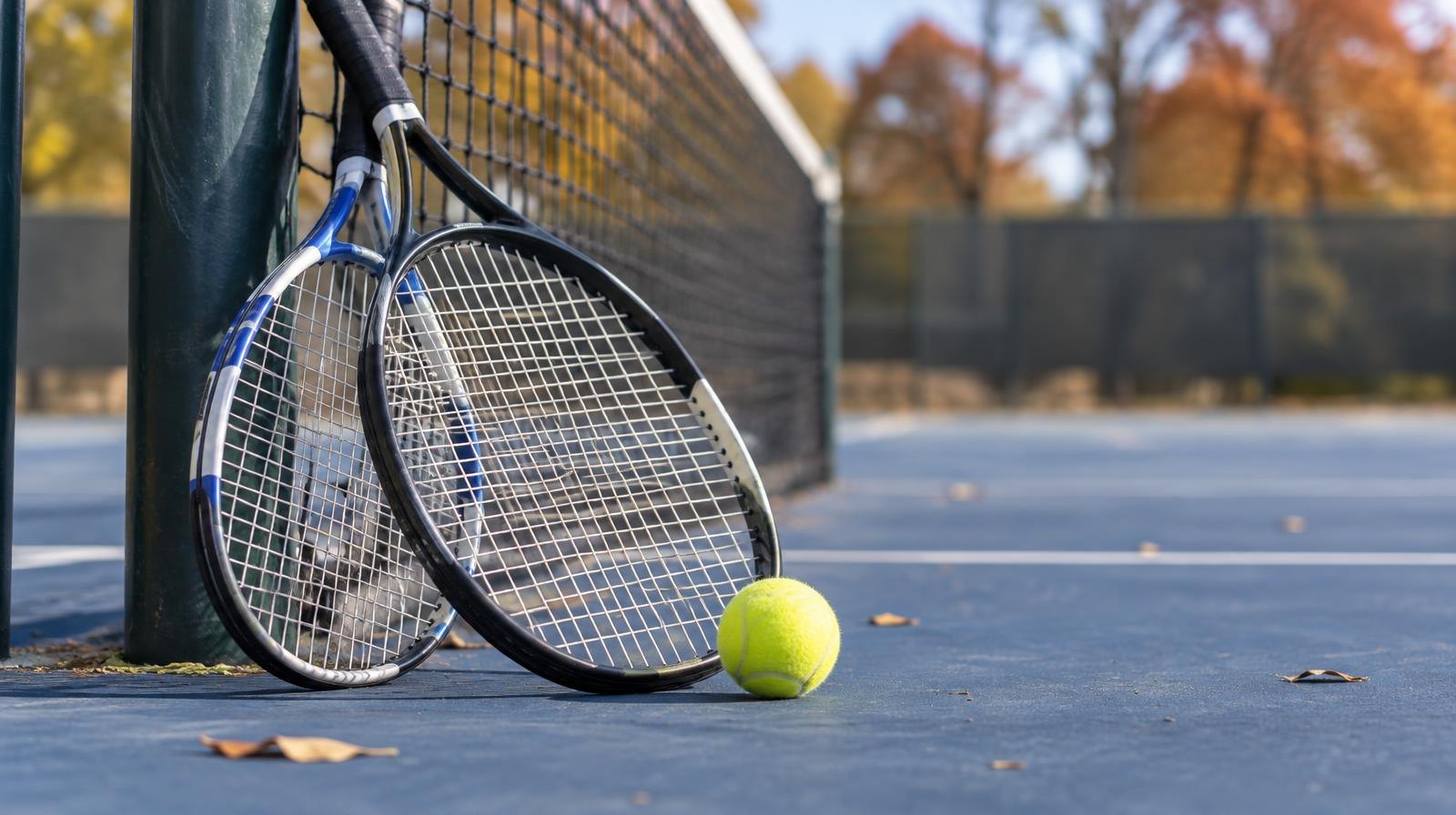Two tennis rackets and a yellow ball resting on a blue hard court near a net post.