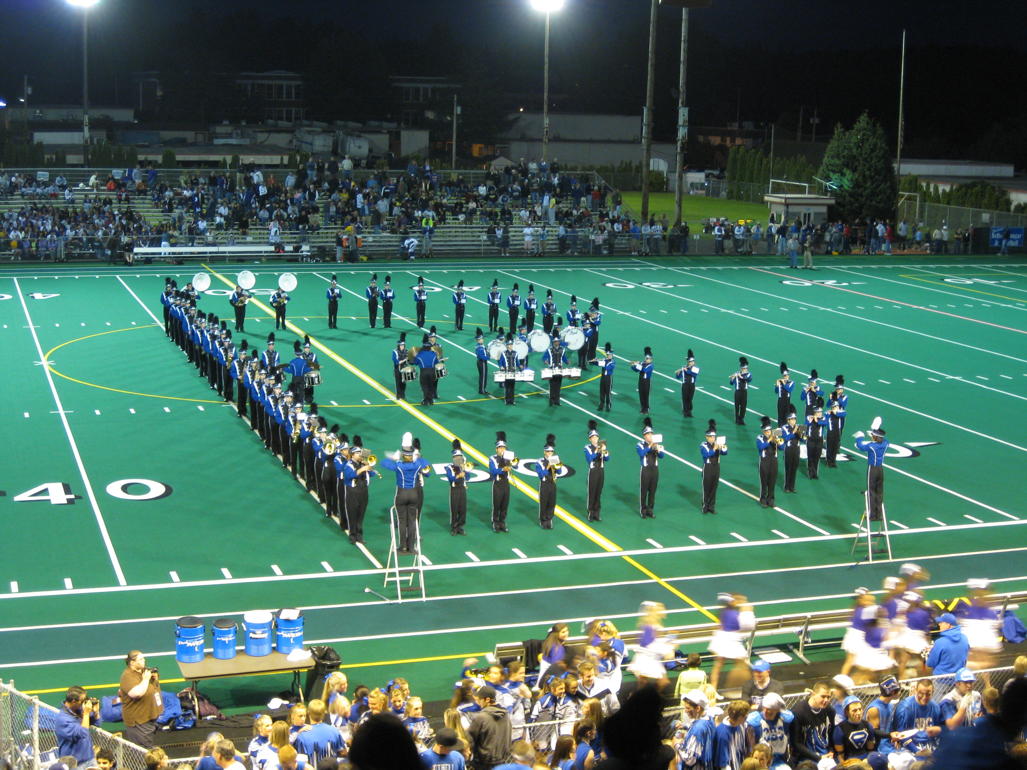 Marching band in blue uniforms performs on a lit football field at night.