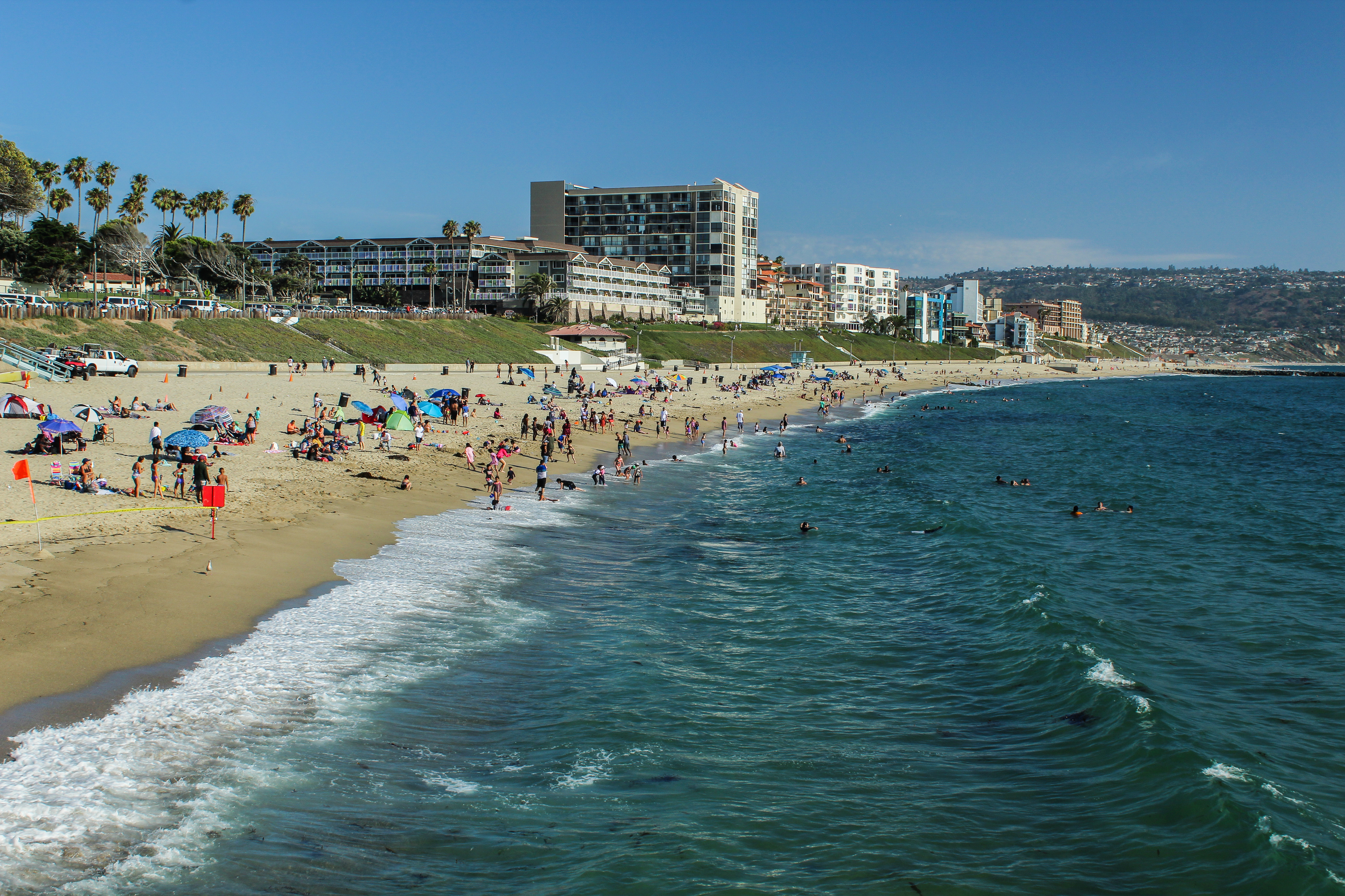 Crowded sandy beach with swimmers in gentle surf and multi-story buildings along the coastline.