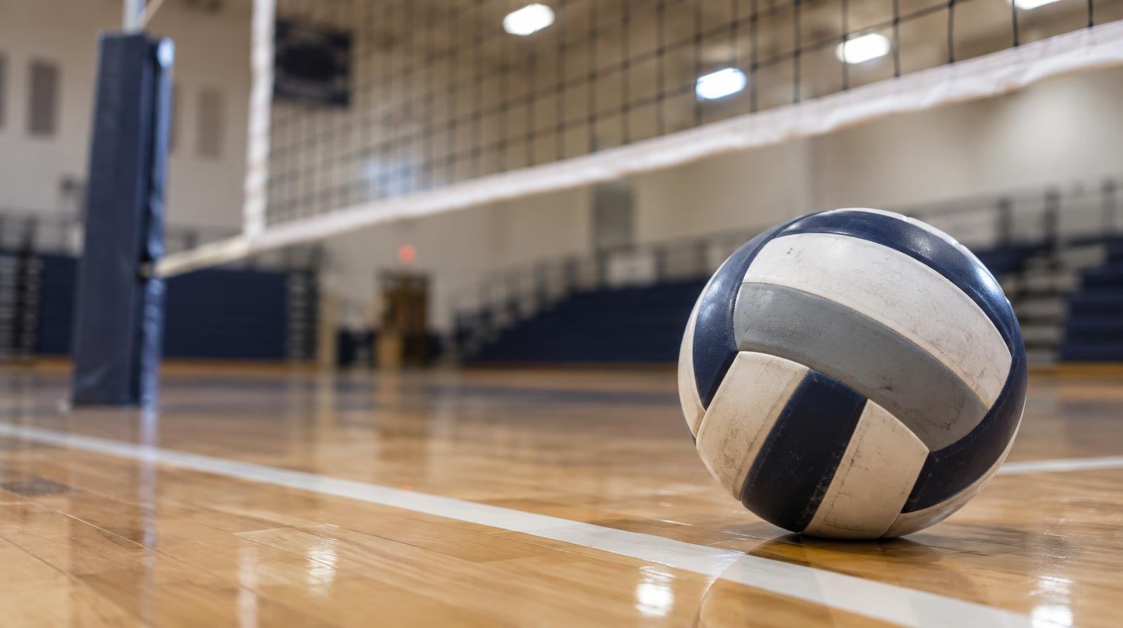 A volleyball resting on a hardwood floor near a volleyball net.