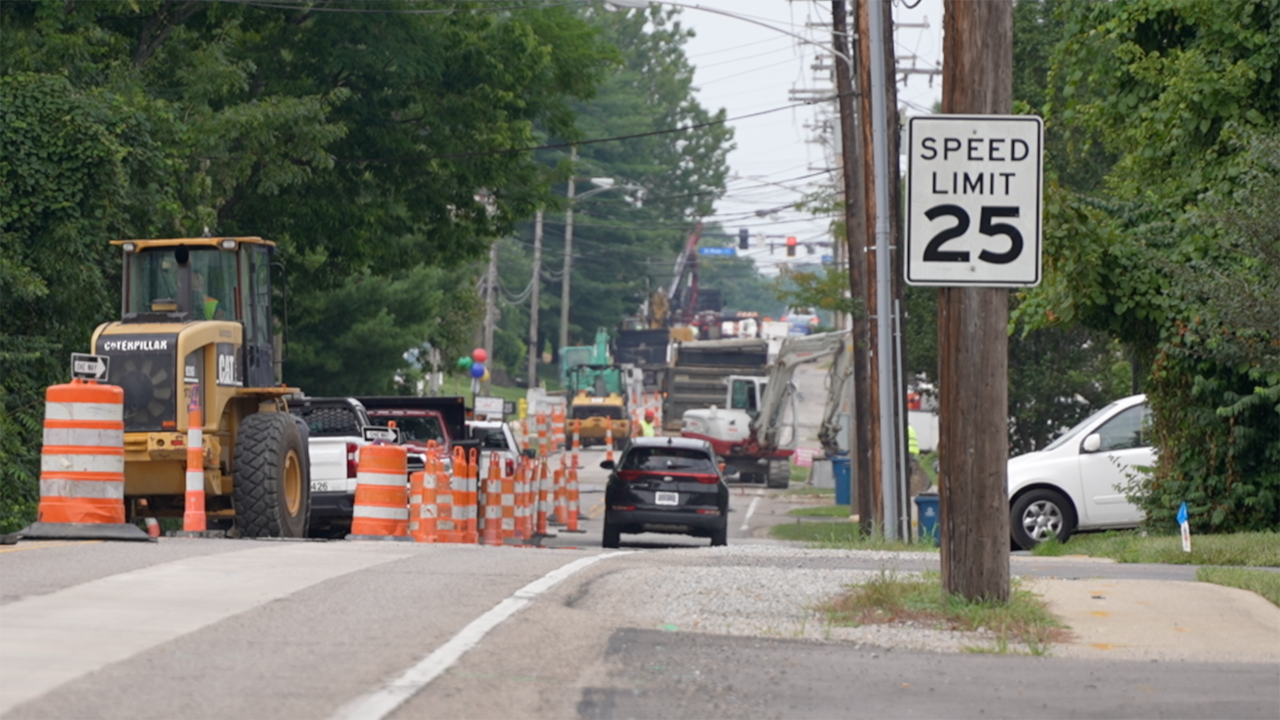 Orange traffic barrels and a front-end loader narrow a tree-lined road to one lane.