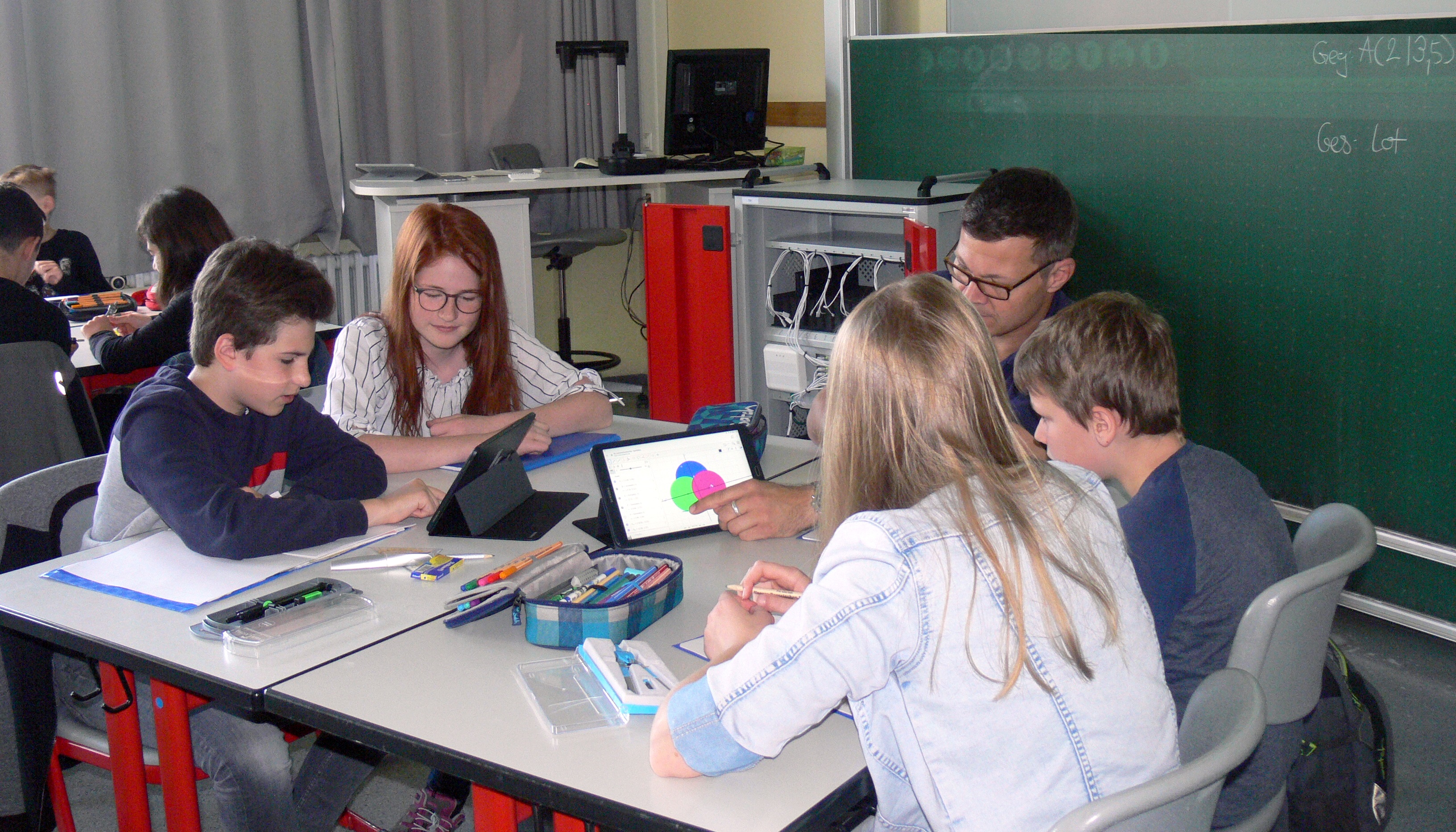 Students and a teacher gathered around a table, one tablet showing a colorful chart.