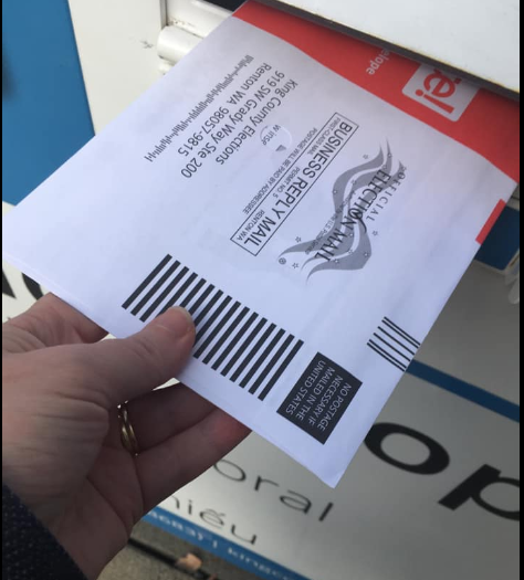 A hand inserting an official election mail envelope into a blue USPS mailbox.