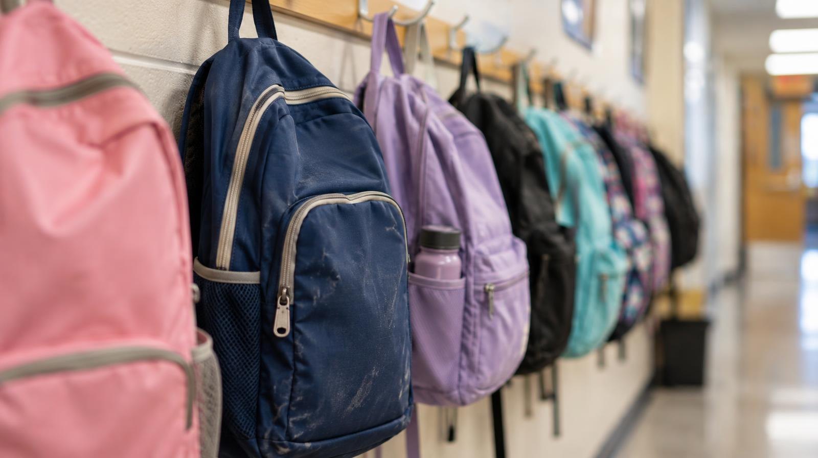 Colorful student backpacks hanging on hooks along a school hallway wall.