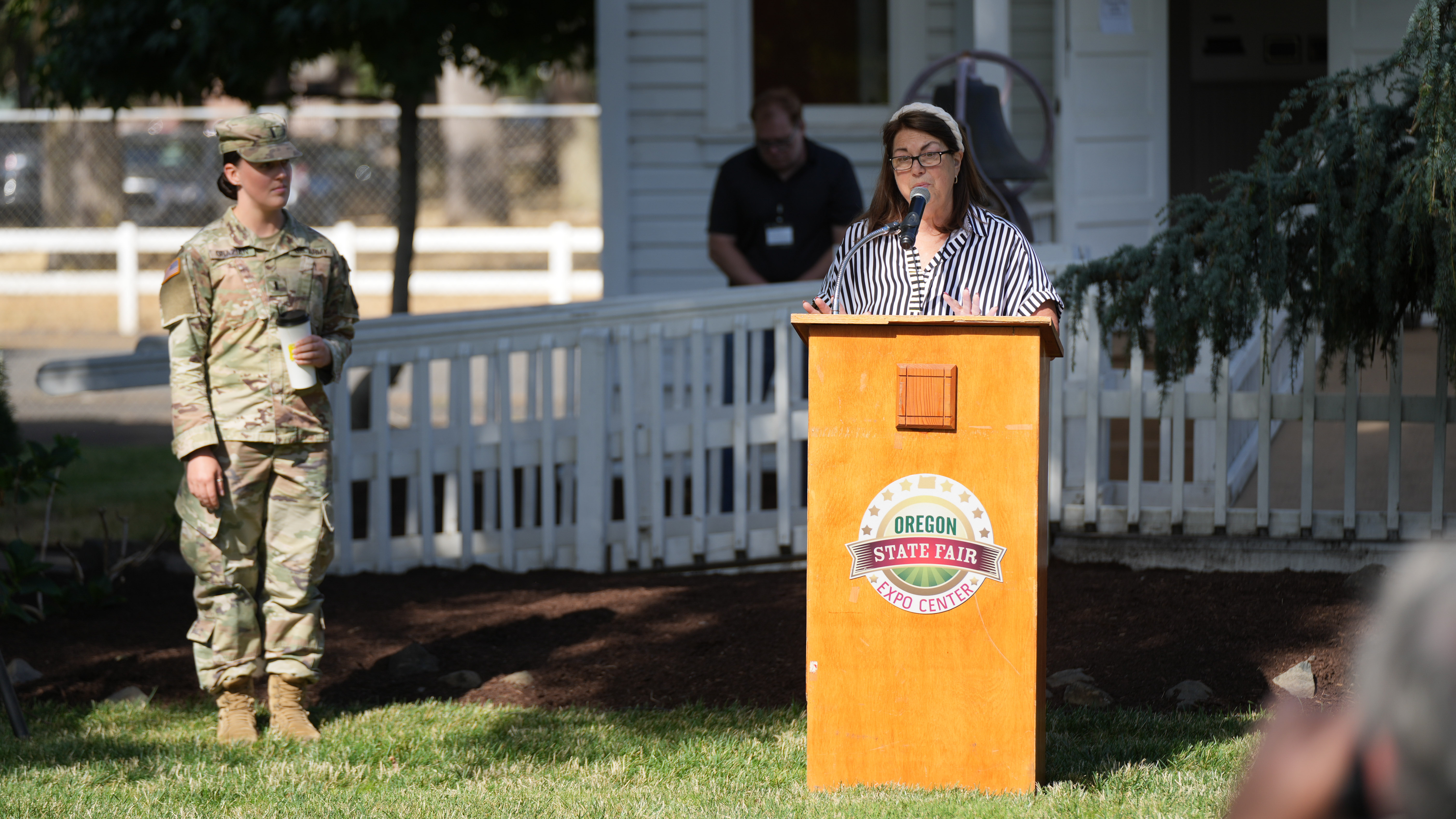 Speaker at an Oregon State Fair podium outdoors, soldier in camouflage uniform standing nearby.