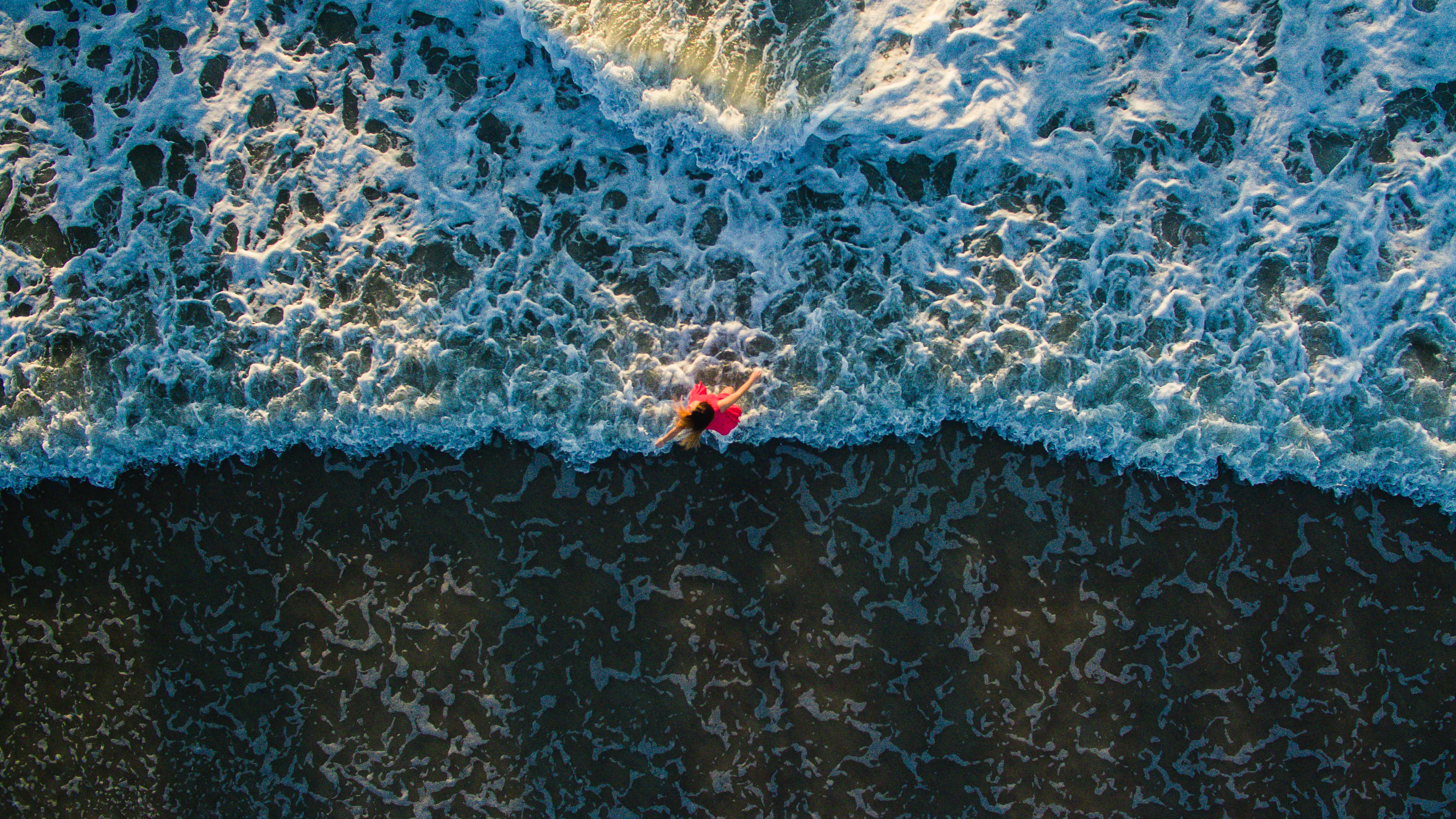 Aerial view of a person in red lying in breaking ocean surf with white foam.