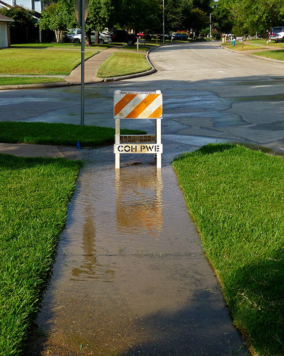 Road-closure barricade standing in brown floodwater on a residential sidewalk.
