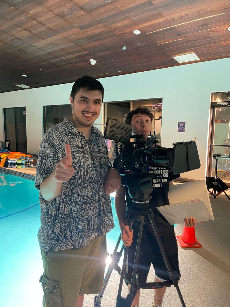 Two young men stand beside a cinema camera on a tripod near an indoor pool.