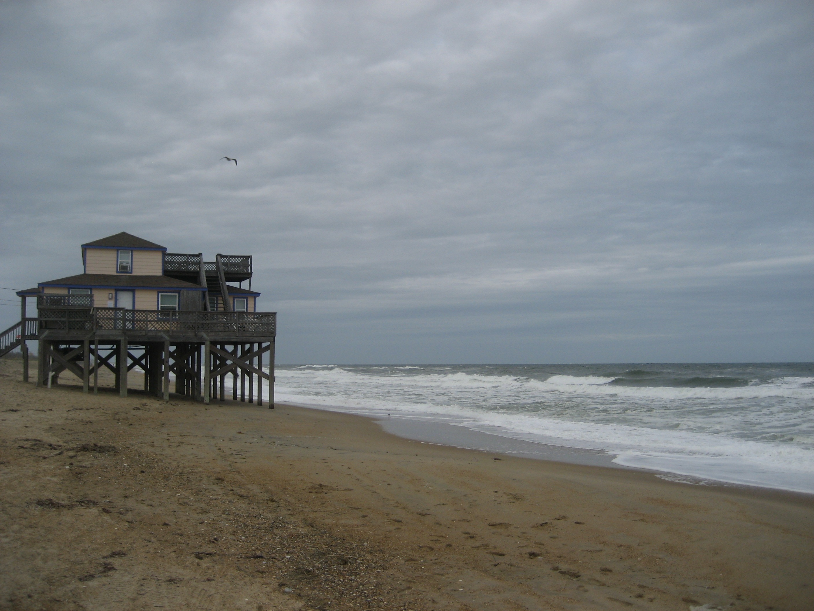 A wooden stilted beach house sits on a sandy shore with ocean waves behind it.