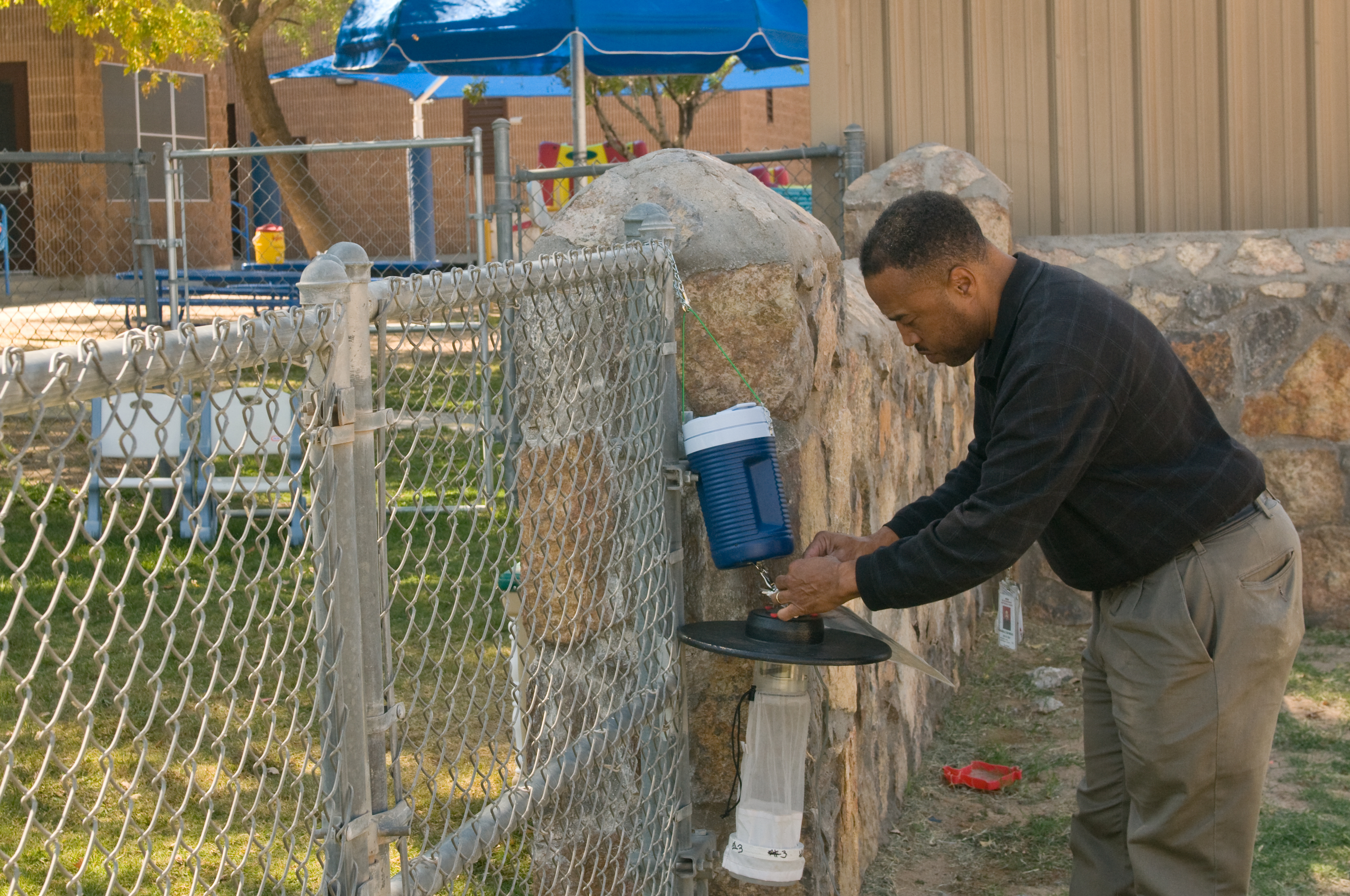 A technician bends over to connect a blue cooler to a mosquito trap mounted on a stone post outdoors.