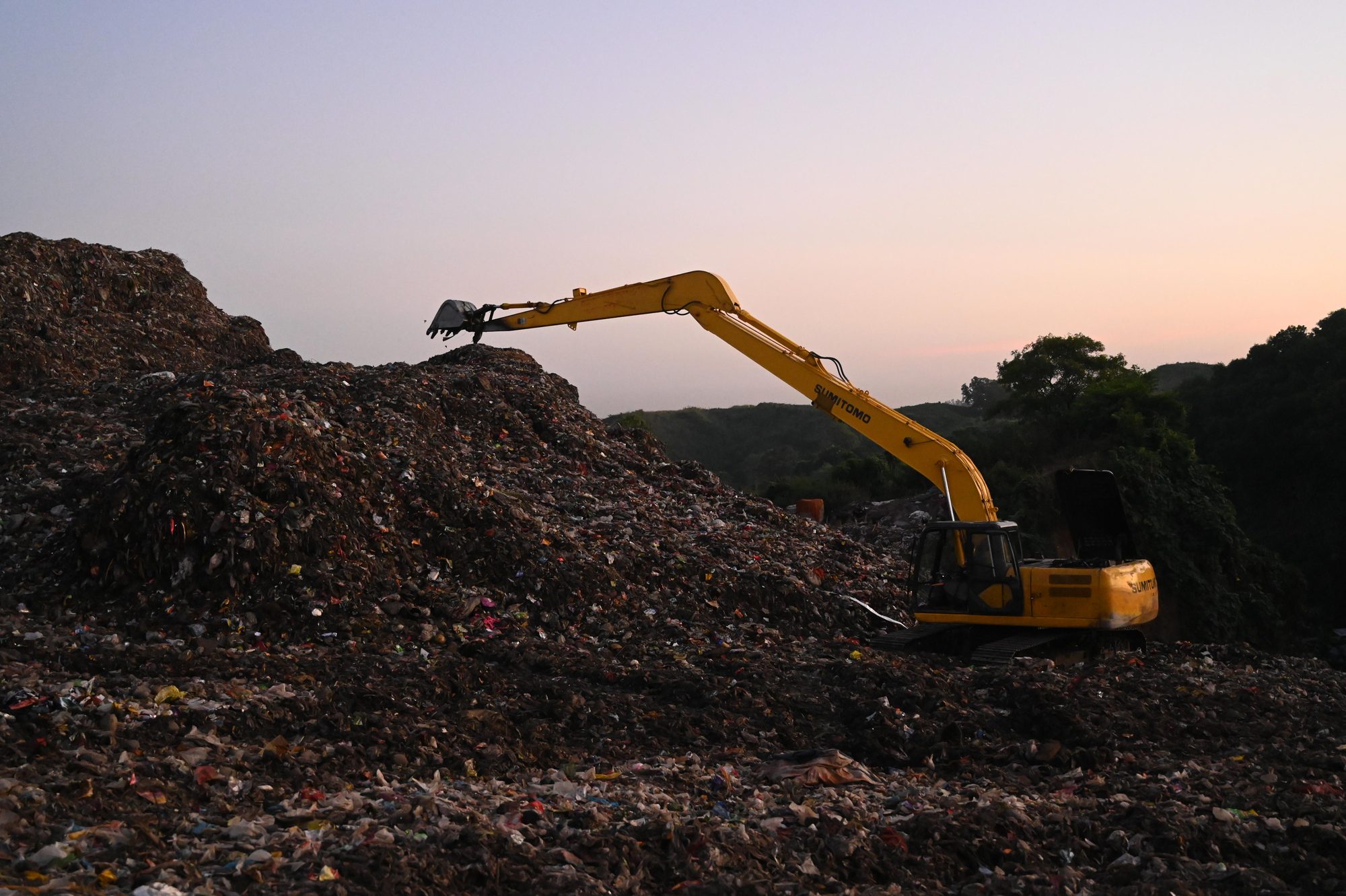 Yellow excavator operates amidst piles of waste in a Bangladesh landfill at sunset.