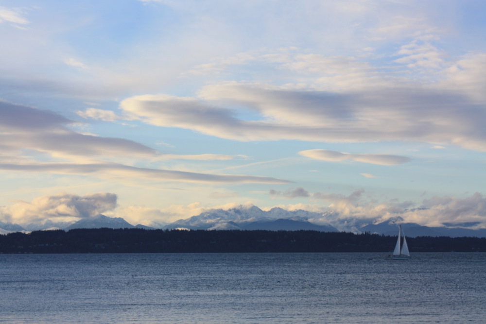 Calm water with a sailboat, forested shoreline, and snow-capped mountains under a partly cloudy sky.