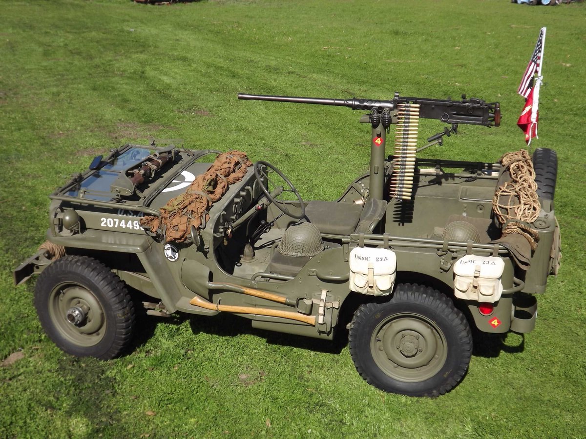 Olive-drab WWII military jeep with mounted machine gun and American flag on grass.