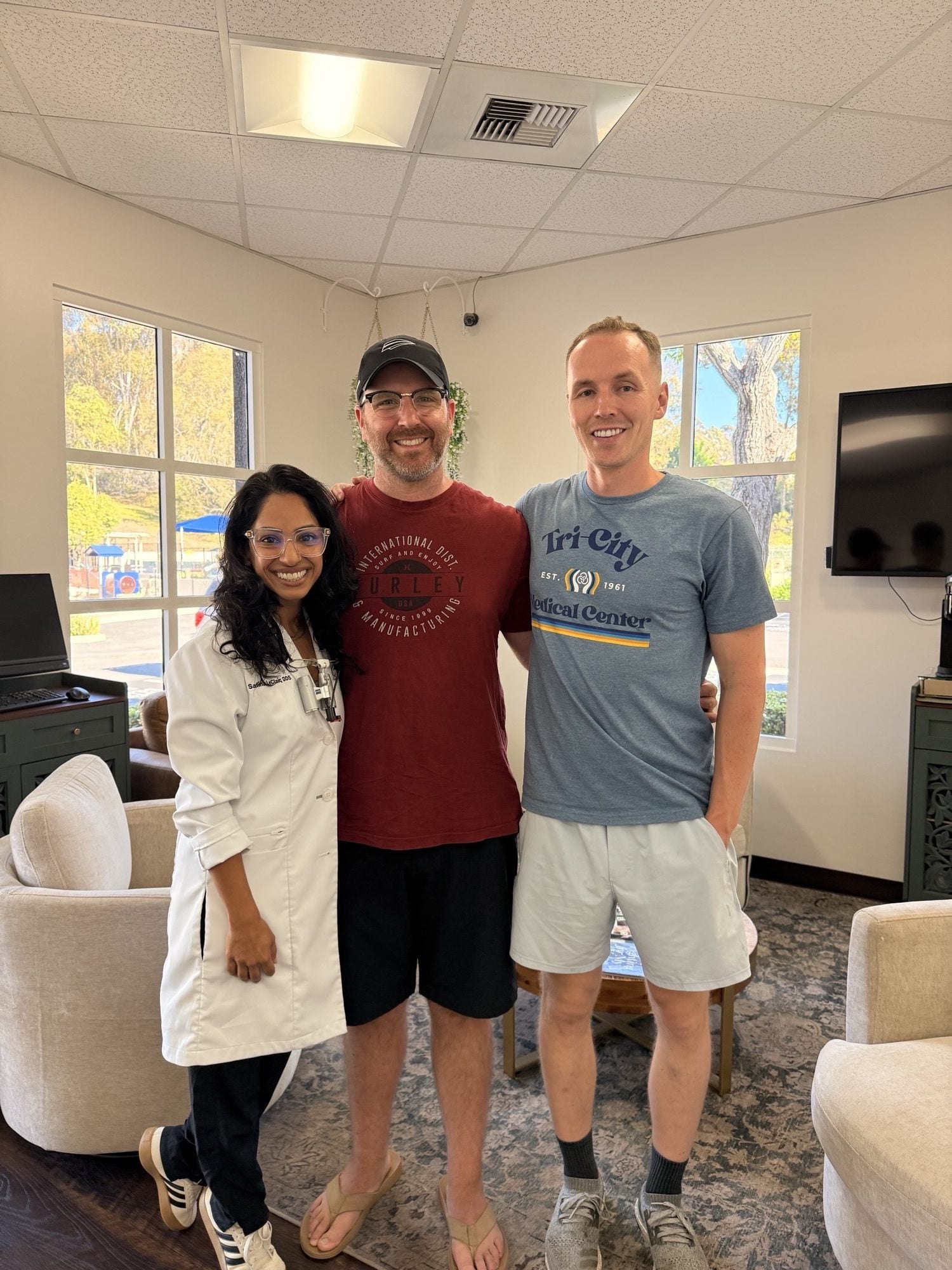 Three people smile together in a bright dental office waiting room, one wearing a white lab coat.