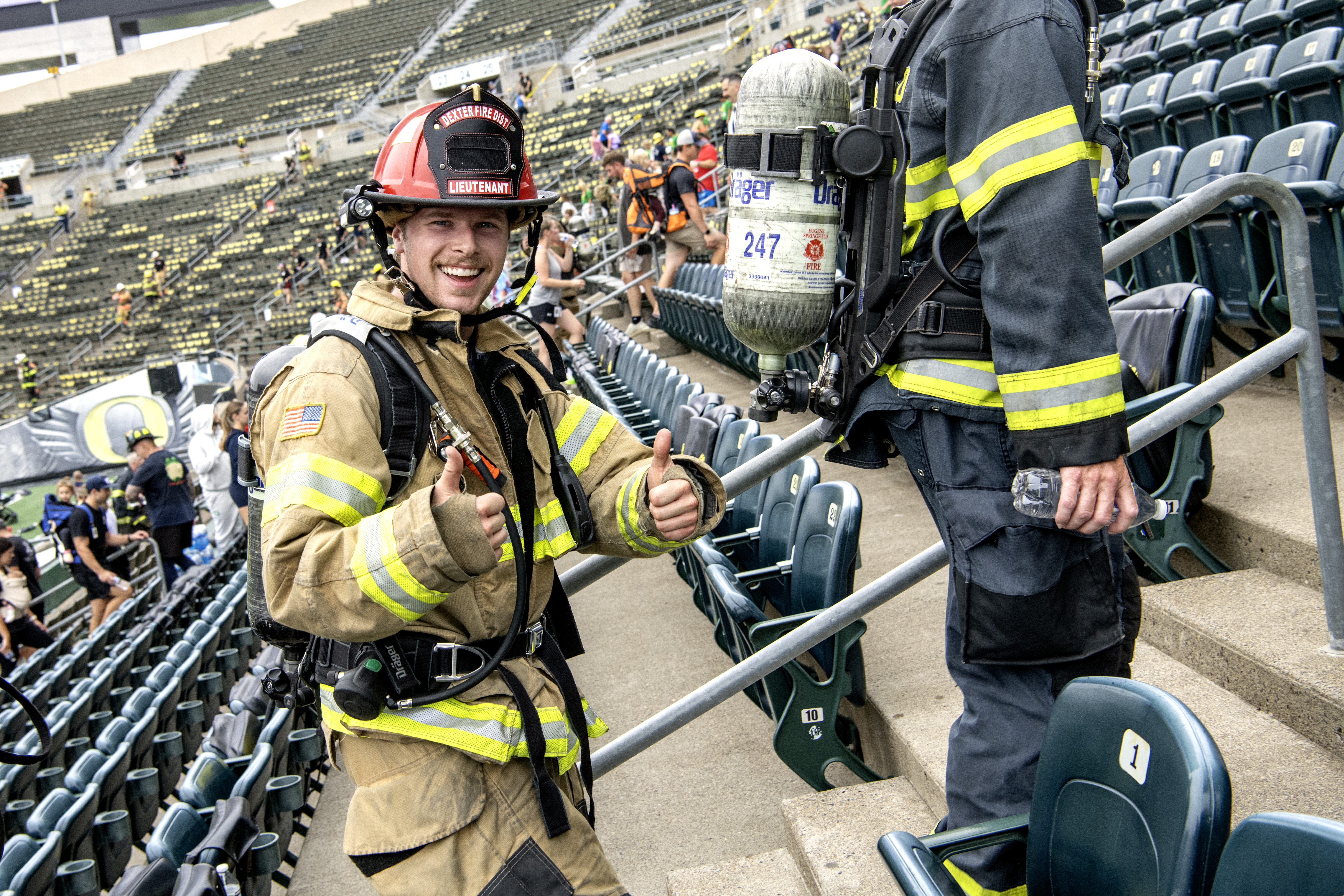 Smiling firefighter in full gear and air tank gives thumbs up while climbing stadium stairs.