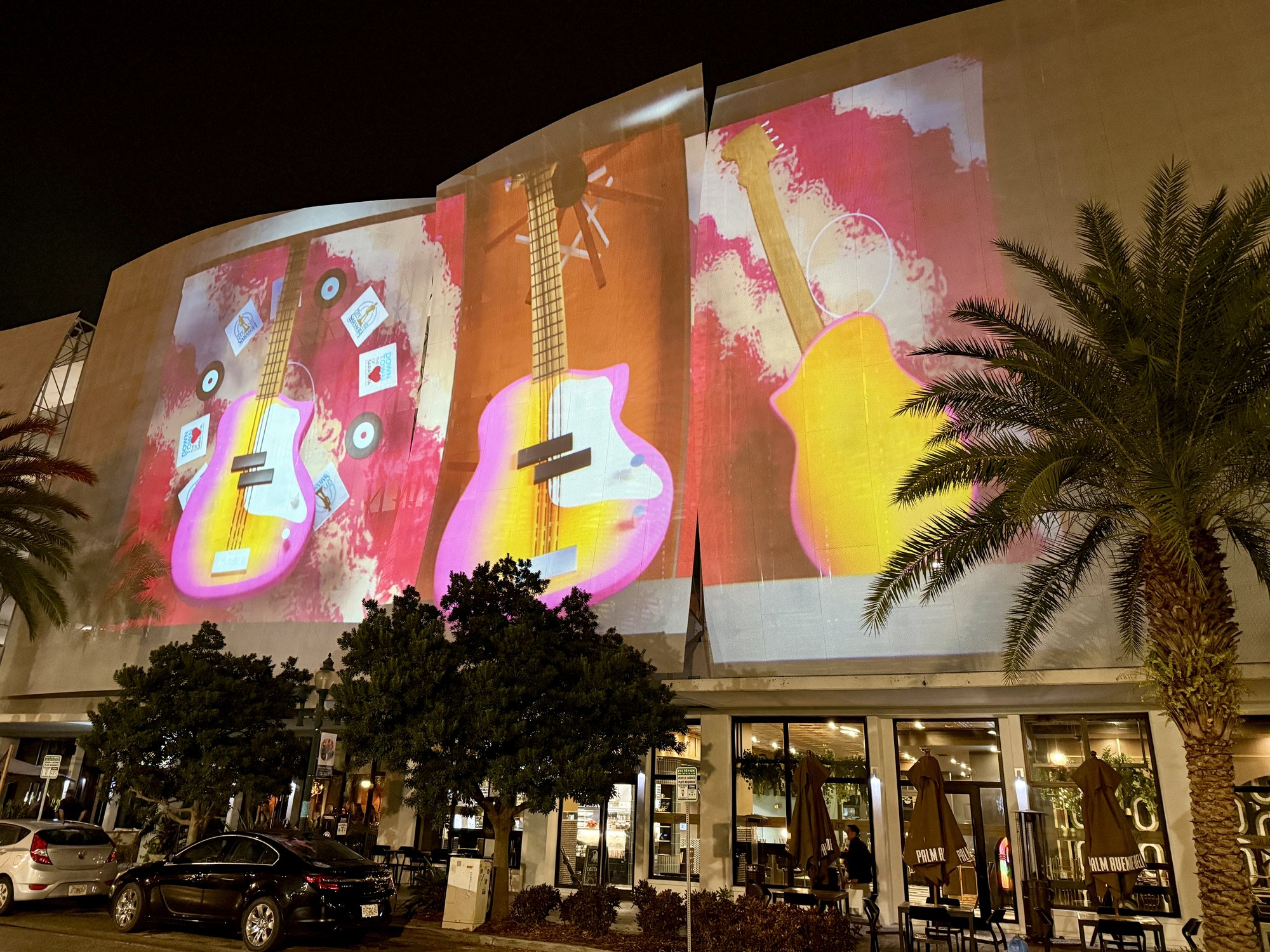 Vivid guitar projections in red and yellow cover a curved building facade at night, flanked by palm trees.