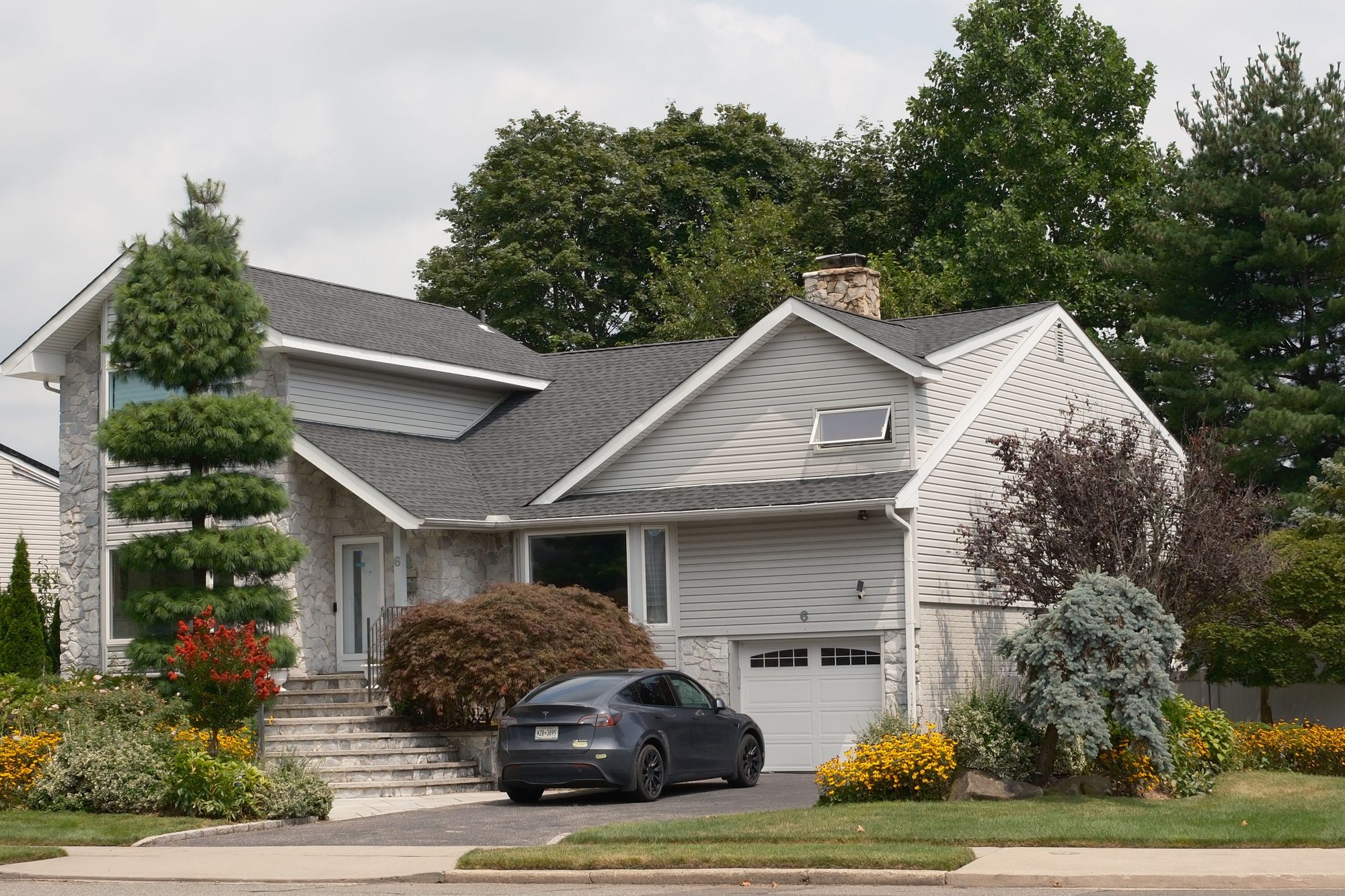 A gray two-story suburban house surrounded by lush greenery with a car parked in the driveway.