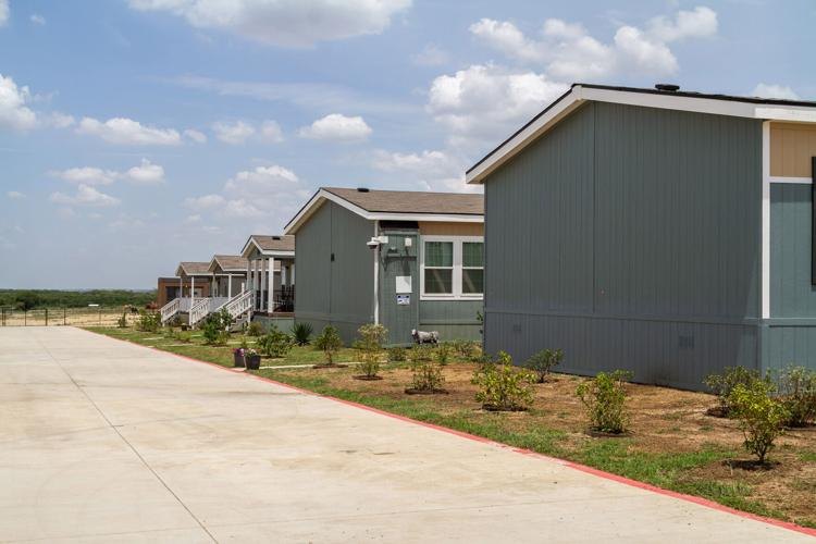 Row of gray single-story residential buildings along a concrete driveway with young landscaping.
