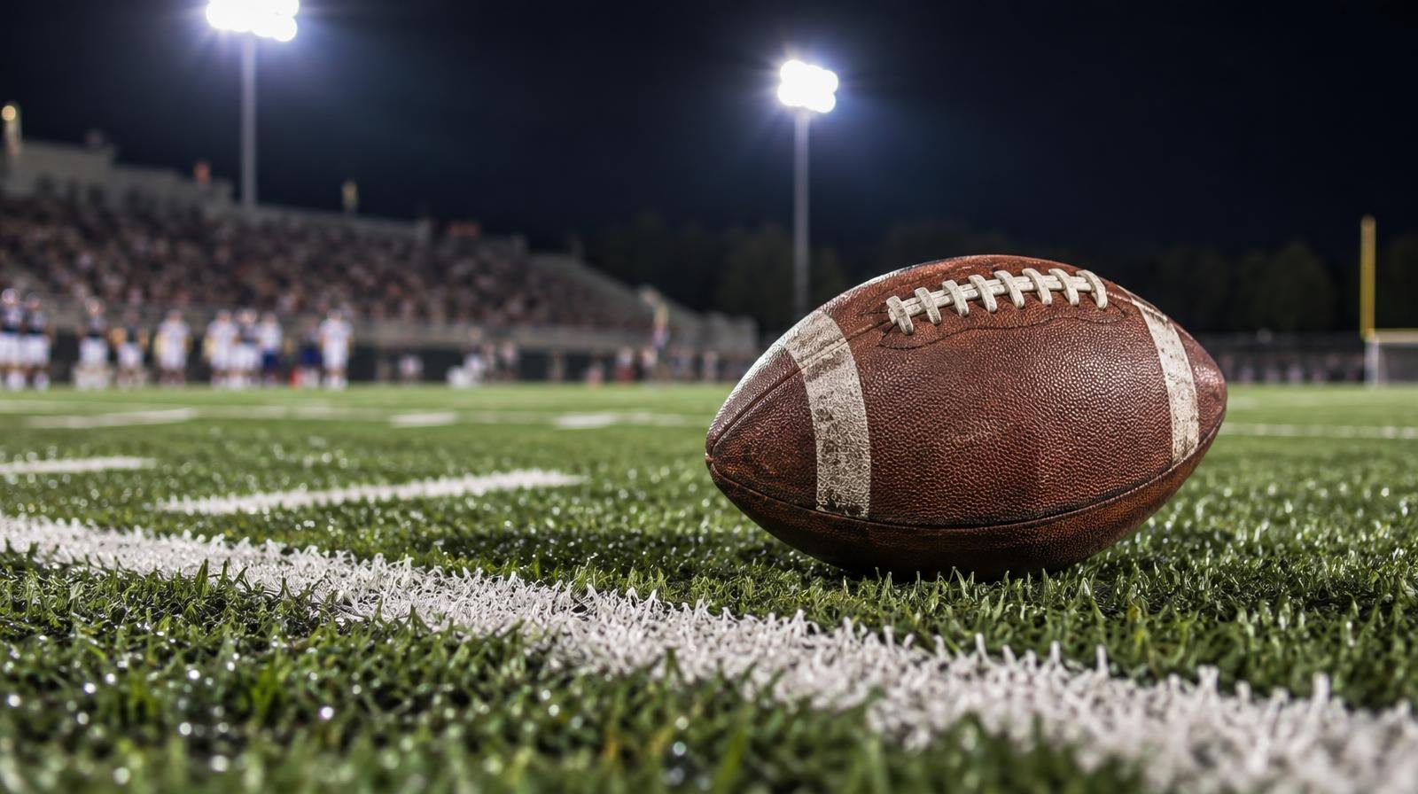 An American football resting on green turf beside a painted white yard line.