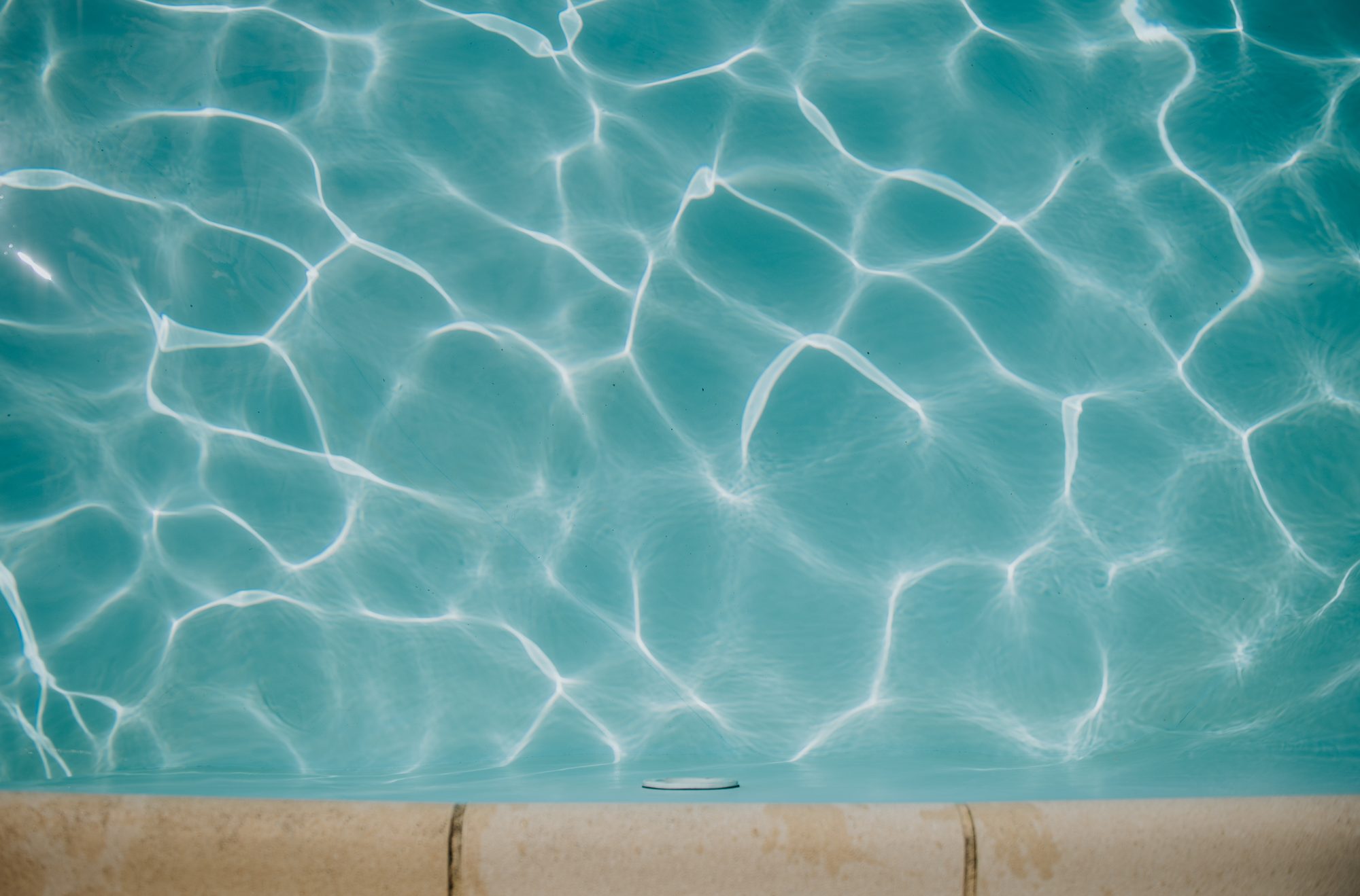 Calm turquoise water with sunlit reflections in a swimming pool surrounded by a light-colored paving surface on a clear day