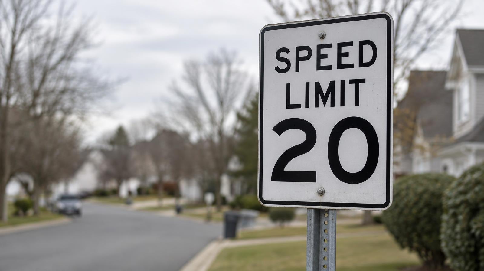A white 20 mph speed limit sign on a metal post along a quiet suburban street.