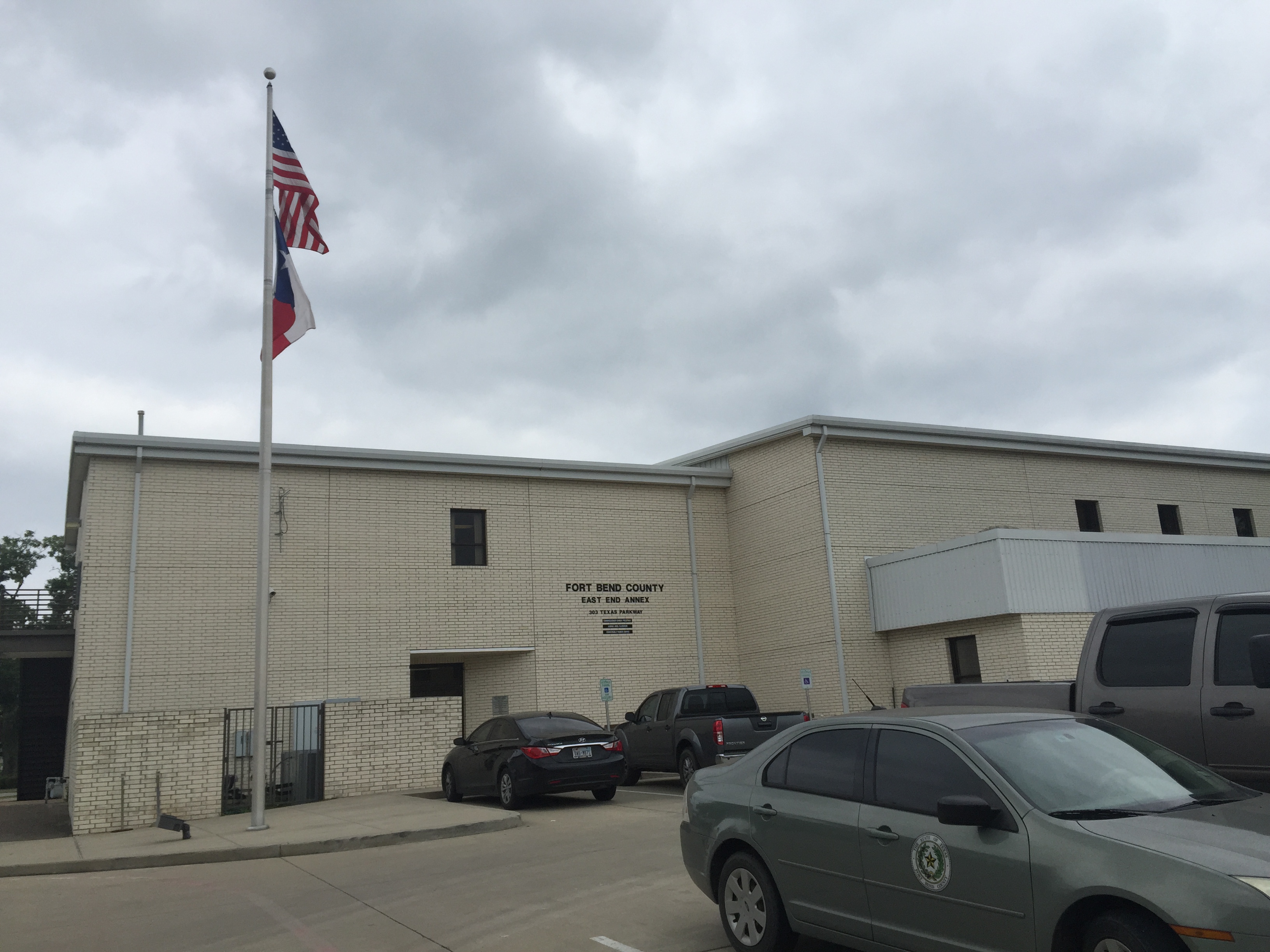 Single-story beige brick government building with American and Texas flags on a flagpole in front.